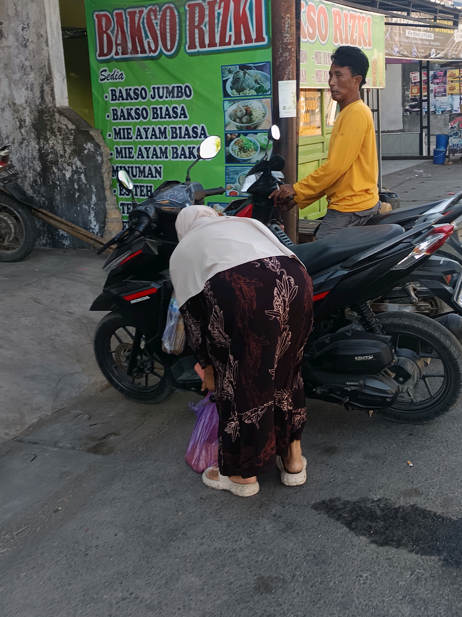 A man in a yellow long-sleeved shirt and grey shorts sits on a black motorcycle, looking to his right. A woman wearing a white hijab and a patterned dark skirt is bent over, reaching for a purple plastic bag near the rear wheel of the motorcycle. The motorcycle is parked on an asphalt road in front of a green banner with the words "BAKSO RIZKI" and pictures of food items. A stone wall is visible to the left of the banner. The scene appears to be on a street in Blega, Indonesia, as indicated by the signage.