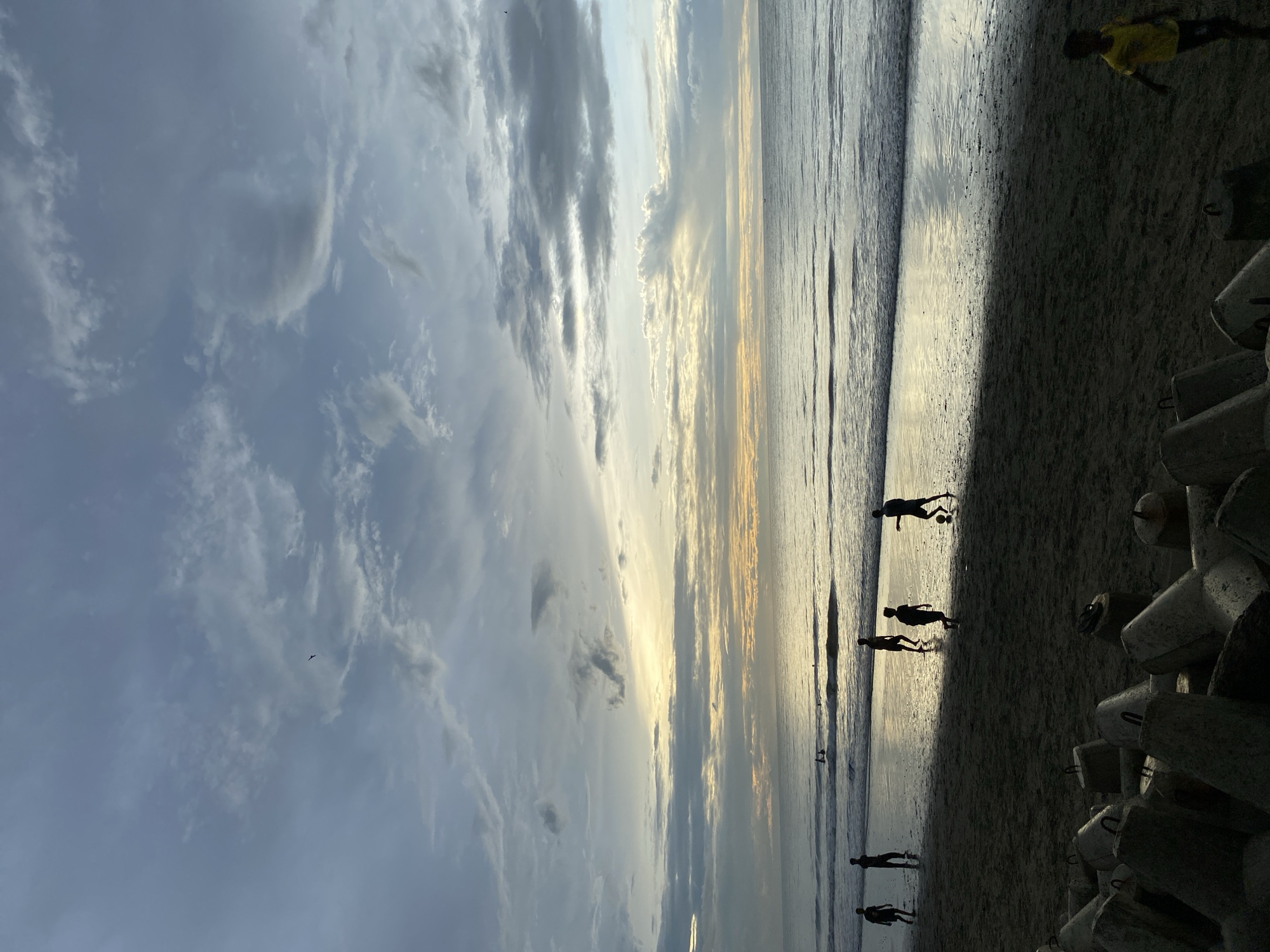 The image captures a beach scene in Bateetimoh, Indonesia, during what appears to be either dawn or dusk, given the soft, diffused light. The sky is filled with a mix of gray and white clouds, with a band of warm, golden light visible on the horizon where the sun is likely low. Gentle waves are rolling onto the sandy shore.
In the mid-ground, several figures are silhouetted against the brighter background, engaged in playing with a ball on the wet sand near the water's edge. These figures appear to be children or young people. Further out in the water, a few other indistinct figures can be seen, possibly swimming or wading.
In the foreground, a collection of large, grey concrete barriers, likely tetrapods or similar coastal defense structures, are arranged on the sand. To the far right of the foreground, a young child in a bright yellow shirt is walking on the sand. The overall atmosphere is peaceful and suggests recreational activity on the beach.
