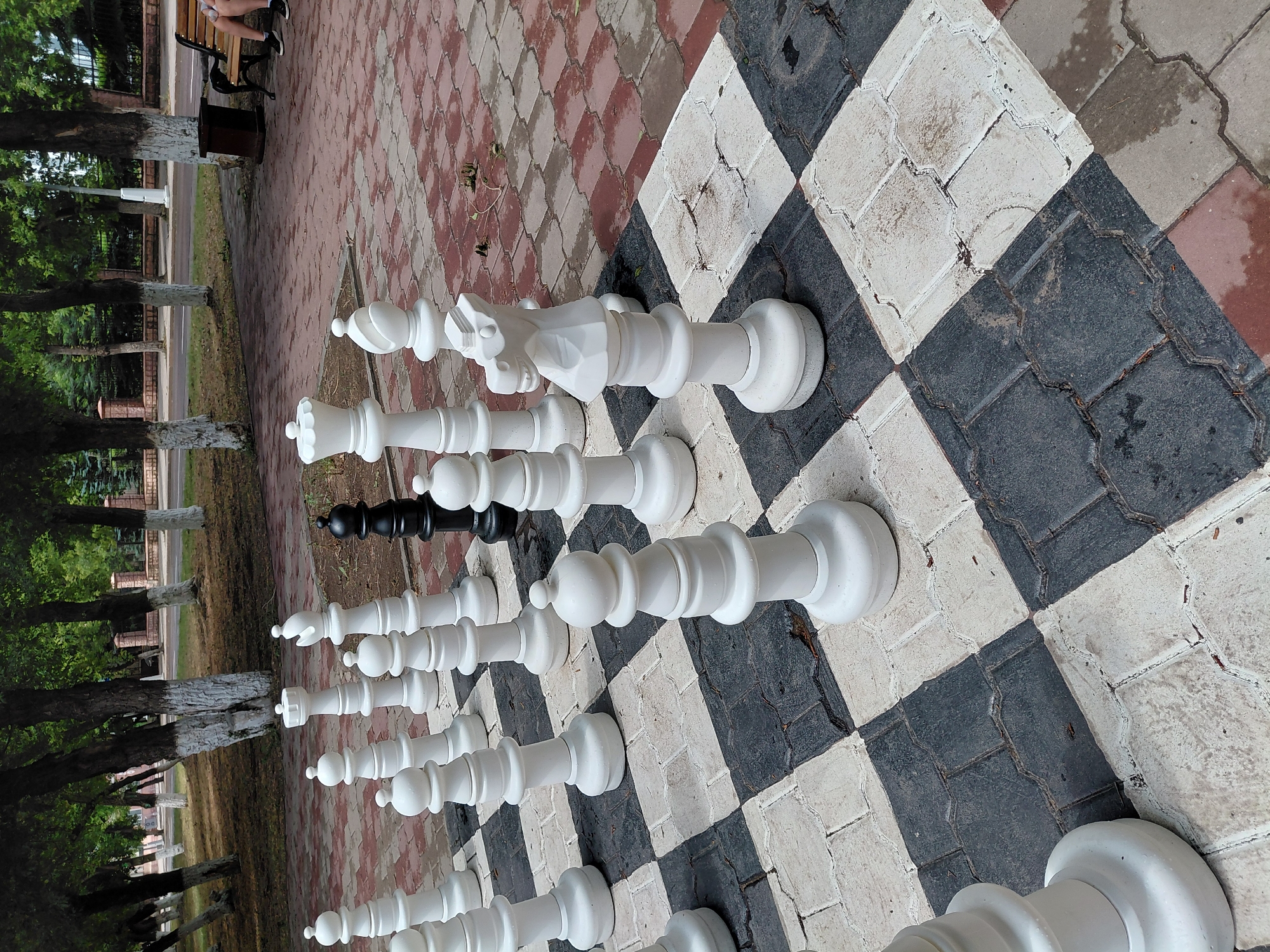 A wide-angle, eye-level shot captures a partially set-up giant chess game on a paved outdoor area, possibly in a park or public square, near trees and a road in Karaganda, Kazakhstan. The chess pieces are predominantly white, with one black rook visible. The chessboard is composed of alternating black and white rectangular pavers. In the background, several people are seated on a wooden bench, and a trash receptacle is nearby. Trees with white-trunked bases line the perimeter of the paved area, and a brick fence is visible further in the distance. The overall scene suggests a public recreational space.