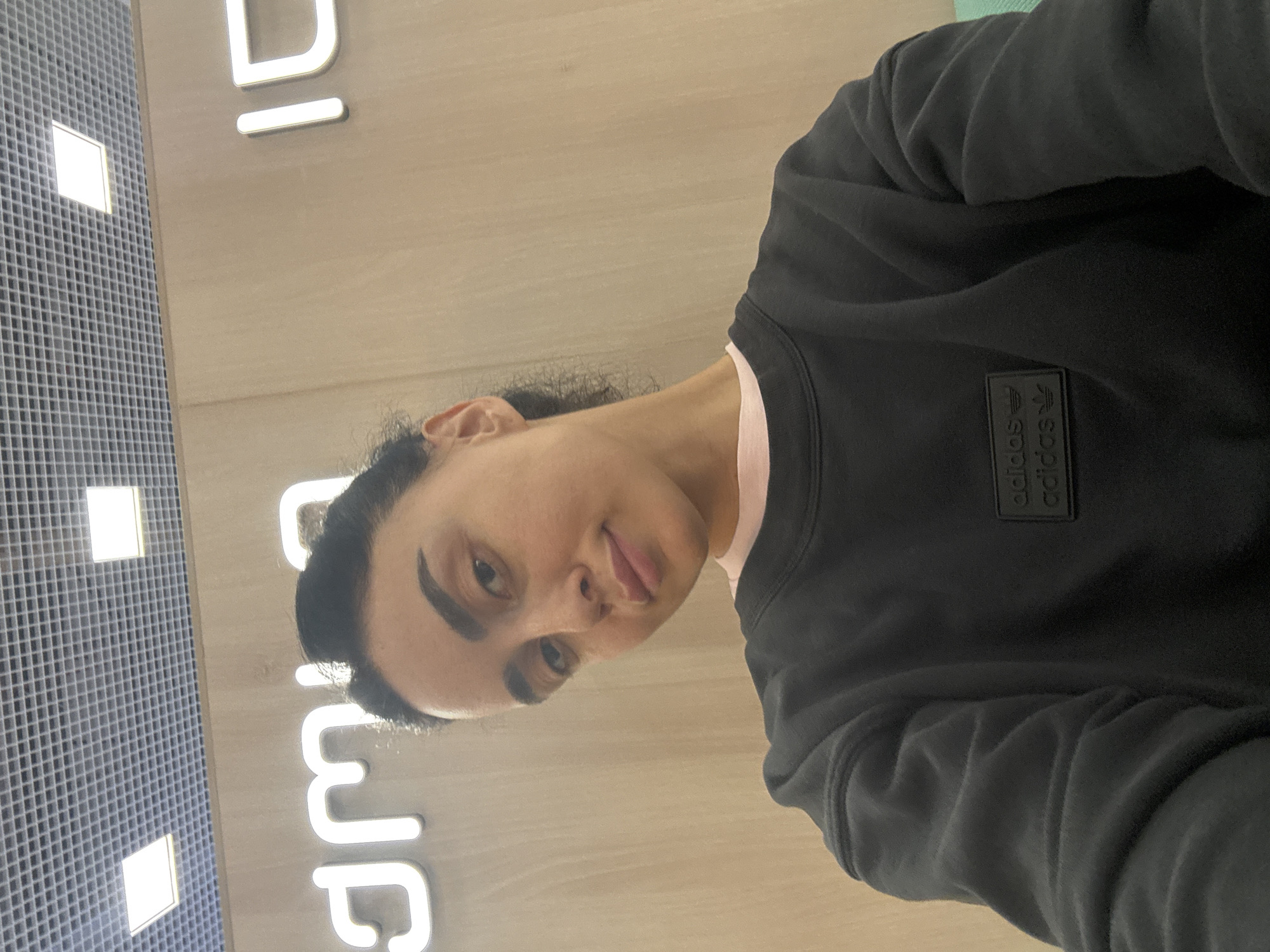 An indoor, close-up selfie shows a person with dark hair, looking directly at the camera with a slight, playful smirk. The image is rotated 90 degrees clockwise, placing the person sideways. They are wearing a black long-sleeved sweatshirt, with a hint of a light pink collar visible underneath. A black, embossed rectangular patch featuring the Adidas logo with the word "adidas" repeated twice, accompanied by the three-stripe motif, is visible on the right side of the sweatshirt.
The background consists of a light-colored, possibly wood-paneled wall or surface, where illuminated letters are mounted. Partial letters "DI" and "mp" are visible, suggesting part of a brand name or sign. To the left, a grid-patterned wall or ceiling panel with bright, square-shaped lights further indicates an indoor commercial or public setting, like a store or showroom. The lighting is artificial, and the time of day is not discernible.