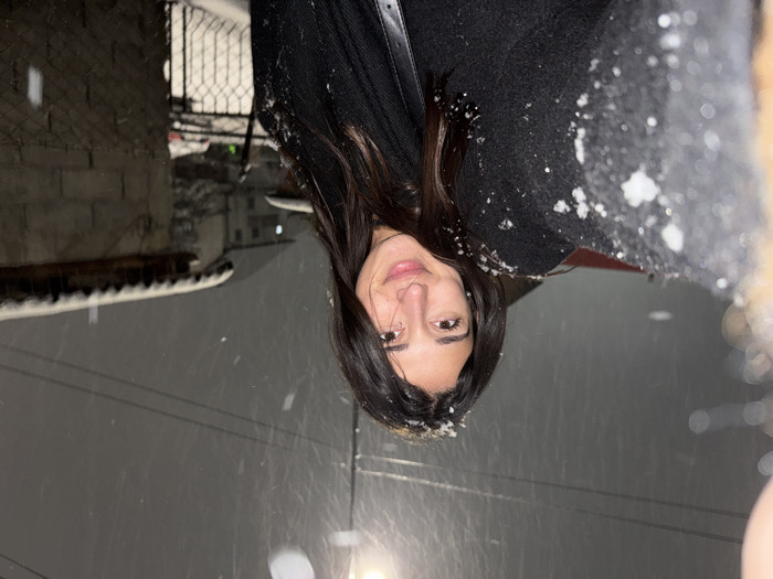 The image features a young woman outdoors at night in Cibukovac, Serbia, during a snowfall. She is positioned centrally, looking directly at the viewer with a slight smile. Her long, dark hair is visible, with delicate snowflakes resting on it and her dark-colored coat or jacket. The ongoing snowfall is evident from the numerous white flakes suspended in the air and accumulated on her clothing.
The background is dimly lit and somewhat blurred, indicating low light conditions and a focus on the subject. Visible elements include what appears to be a building with a textured wall and a metal fence or gate on the left side. Utility wires or power lines traverse the middle distance. A snow-covered ledge or roof edge is also discernible in the background, reinforcing the wintry scene. The overall ambiance suggests a cold, snowy night. The woman's expression implies contentment or enjoyment despite the weather. There is no visible text in the image.