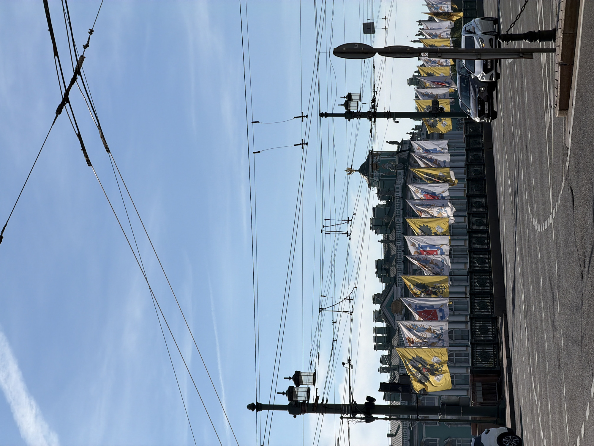 The image captures a view from a street in Saint Petersburg, Russia, likely overlooking the Neva River, given the presence of flags and architectural style. In the foreground, a paved road with white lane markings is visible. A white car is parked on the left side of the road, and further ahead, two other cars are driving on the right side of the road.
Dominating the mid-ground is a line of large, ornate flags, predominantly yellow and white, displayed on poles along a bridge or embankment. These flags feature intricate emblems, suggesting a ceremonial or patriotic occasion. Behind the flags, a grand building with a green facade and numerous arched windows can be seen. The building is adorned with sculptures and features multiple chimneys or decorative elements along its roofline. A golden eagle sculpture is prominent atop a dome on the building.
Overhead, a complex network of electrical wires and trolleybus lines crisscrosses the sky. Several lampposts, including one on the left with two lanterns, are positioned along the street. The sky is a clear blue with a few wispy white clouds, indicating a bright, sunny day. The overall scene conveys a sense of urban grandeur and public celebration.