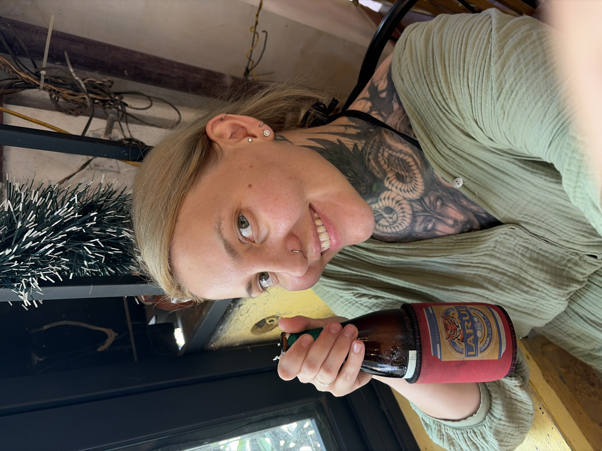 A young woman with blonde hair and fair skin is captured in a close-up, smiling directly at the camera while holding a beer bottle. She has multiple ear piercings, including a cluster stud on her lobe and a silver hoop nose ring. Prominent blackwork tattoos, featuring intricate designs resembling snakes or mythical creatures, cover her neck and extend onto her shoulder and arm. She is wearing a light green, textured, long-sleeved shirt or dress.
The beer bottle she holds is wrapped in a red koozie, with the brand name "LARUE" clearly visible in yellow and blue, along with a tiger graphic. Faint text, likely "D'EPUI..." (possibly "Depuis"), can also be seen on the label.
The background shows a somewhat cluttered indoor or semi-covered environment. Above her, there are bundled wires, some secured with zip ties, and a wooden beam. To her left, part of a white and green tinsel-like garland is visible. Below, a dark surface, possibly a window or table, can be seen. The natural lighting suggests it's daytime. The woman's relaxed smile conveys a sense of happiness and ease. Given the visible "LARUE" beer, a popular brand in Vietnam, and the provided location context, the scene is set in Hội An Ward, Vietnam.