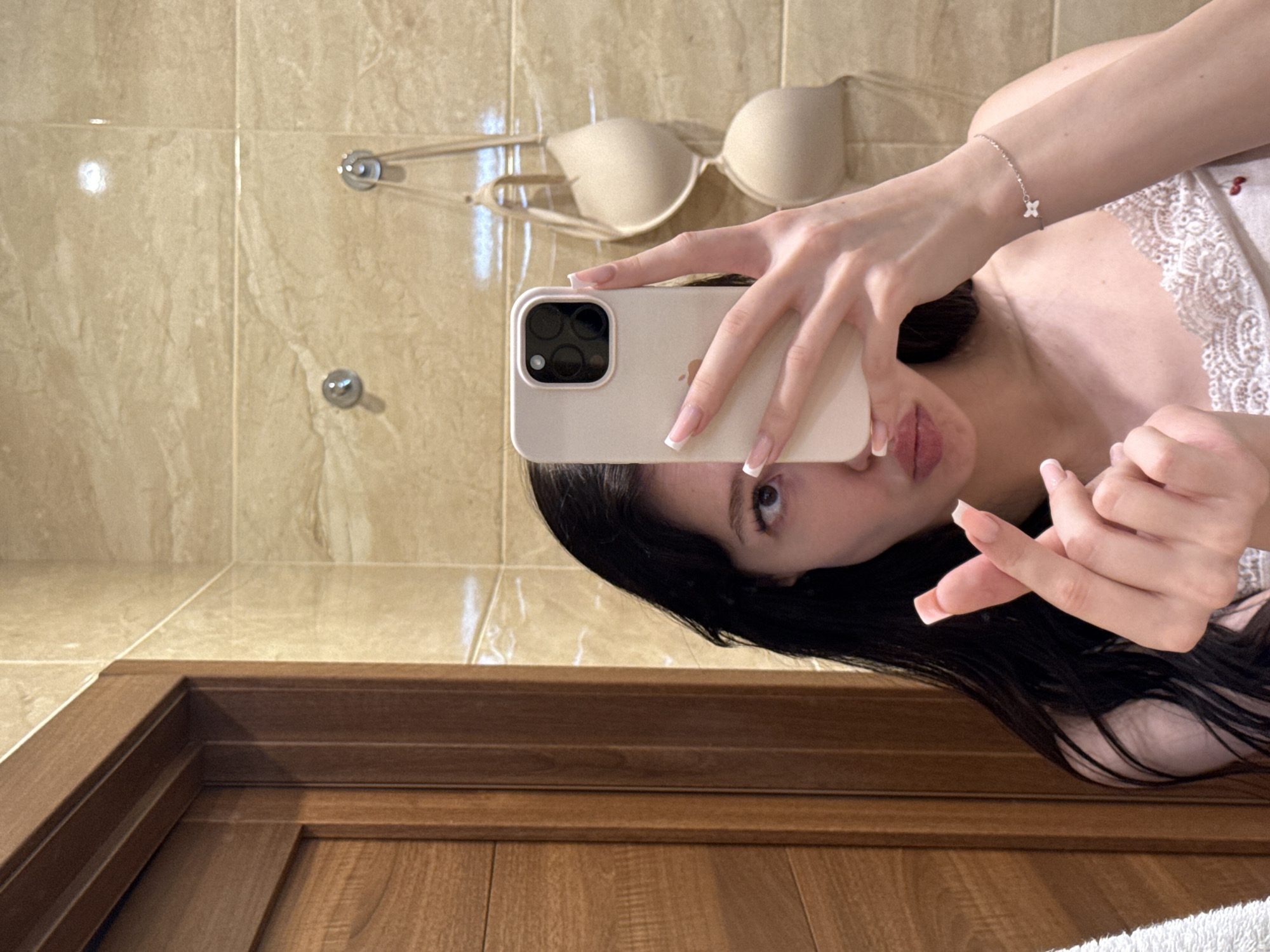 A young woman is taking a mirror selfie in a bathroom. She has long dark hair and is wearing a light-colored top with lace trim. She is holding a smartphone in her right hand, angled to capture her reflection. Her left hand is raised, forming a heart shape with her thumb and index finger. Behind her, a beige bra is hanging on hooks on a tiled wall. The bathroom features light brown wooden door trim. The scene appears to be in a residential setting, possibly an apartment in Khimki, Russia.