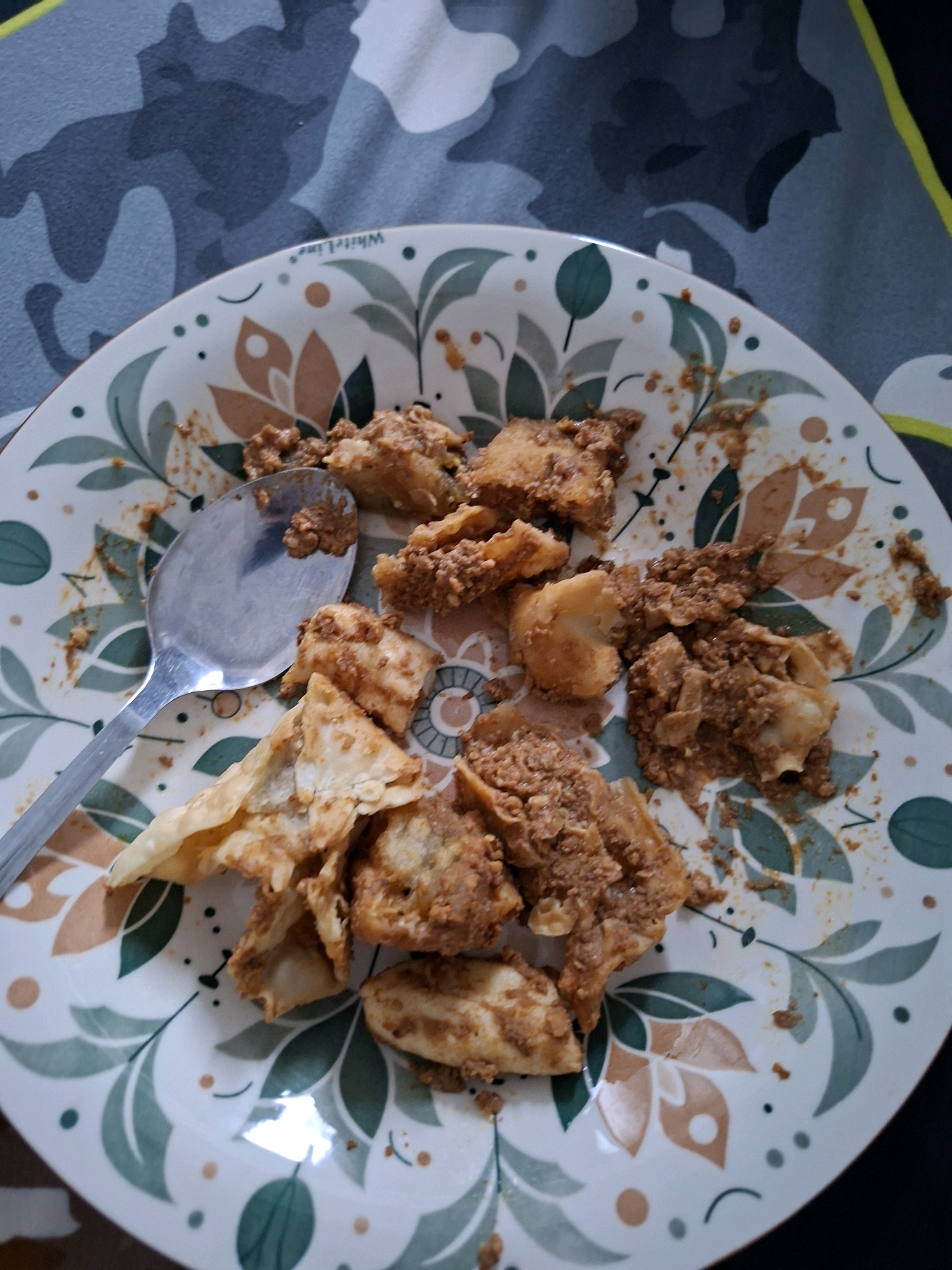 A close-up, overhead shot captures a white plate adorned with a green and brown leaf pattern, filled with Indonesian food. The food consists of several pieces of what appears to be fried dough, some filled with a savory minced meat sauce. A silver spoon rests on the plate, its surface reflecting the light and also bearing a small amount of the food. The plate sits on a surface with a dark grey and black camouflage pattern. The scene is likely indoors, given the lighting and context. The image does not provide enough information to determine the specific location within Sampora, Indonesia.