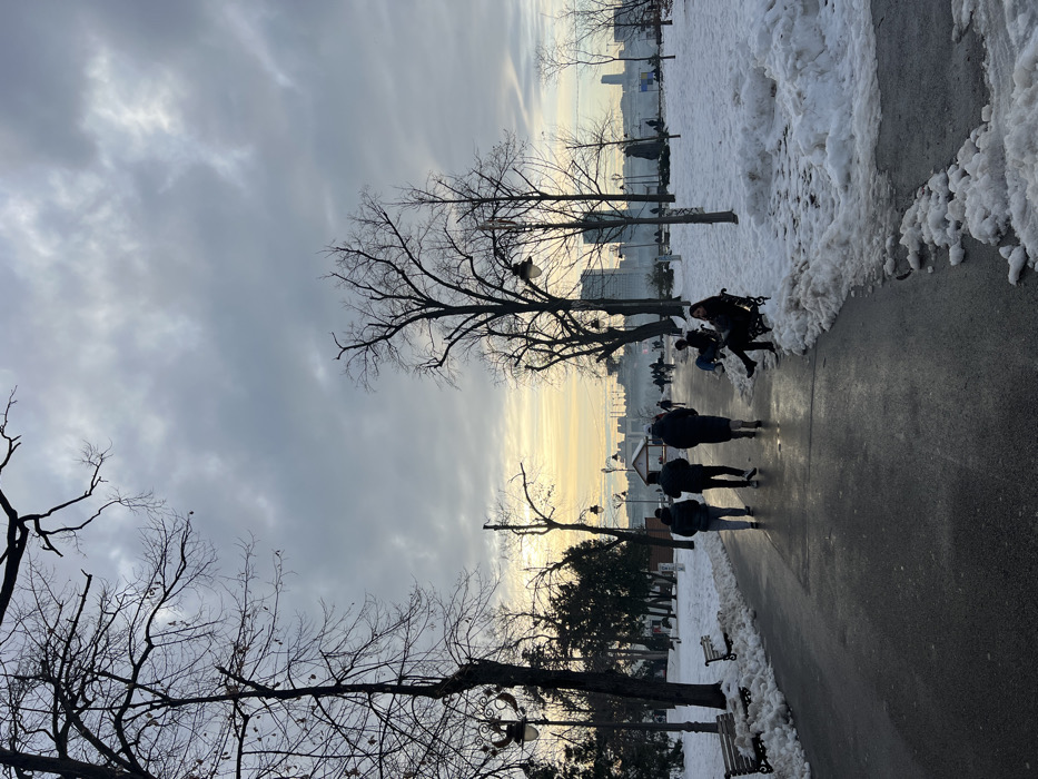 The image displays a winter park scene within Kalemegdan Park, Belgrade, Serbia, under a mostly overcast sky with a warm, yellowish glow on the horizon. A dark, wet-appearing paved pathway, cleared of snow, extends from the foreground towards the background, gently curving to the left. Accumulations of dirty, melting snow are present on both sides of the path and across the park's unpaved areas. Numerous bare deciduous trees are silhouetted against the sky, alongside some evergreen trees on the left. Several ornate dark-colored lampposts with illuminated or reflective fixtures are visible along the path. In the foreground-left, three individuals wearing dark winter coats walk away from the camera along the path. In the foreground-right, one person, dressed in dark clothing, is seated on a snow-dusted park bench, facing partially left. Further into the mid-ground, a small white kiosk-like structure is visible near the path. The background reveals a distant city skyline composed of various buildings, consistent with a view across the Sava or Danube river from Kalemegdan. Multiple other people are dispersed throughout the park in the mid-ground and background, walking or seated on benches.