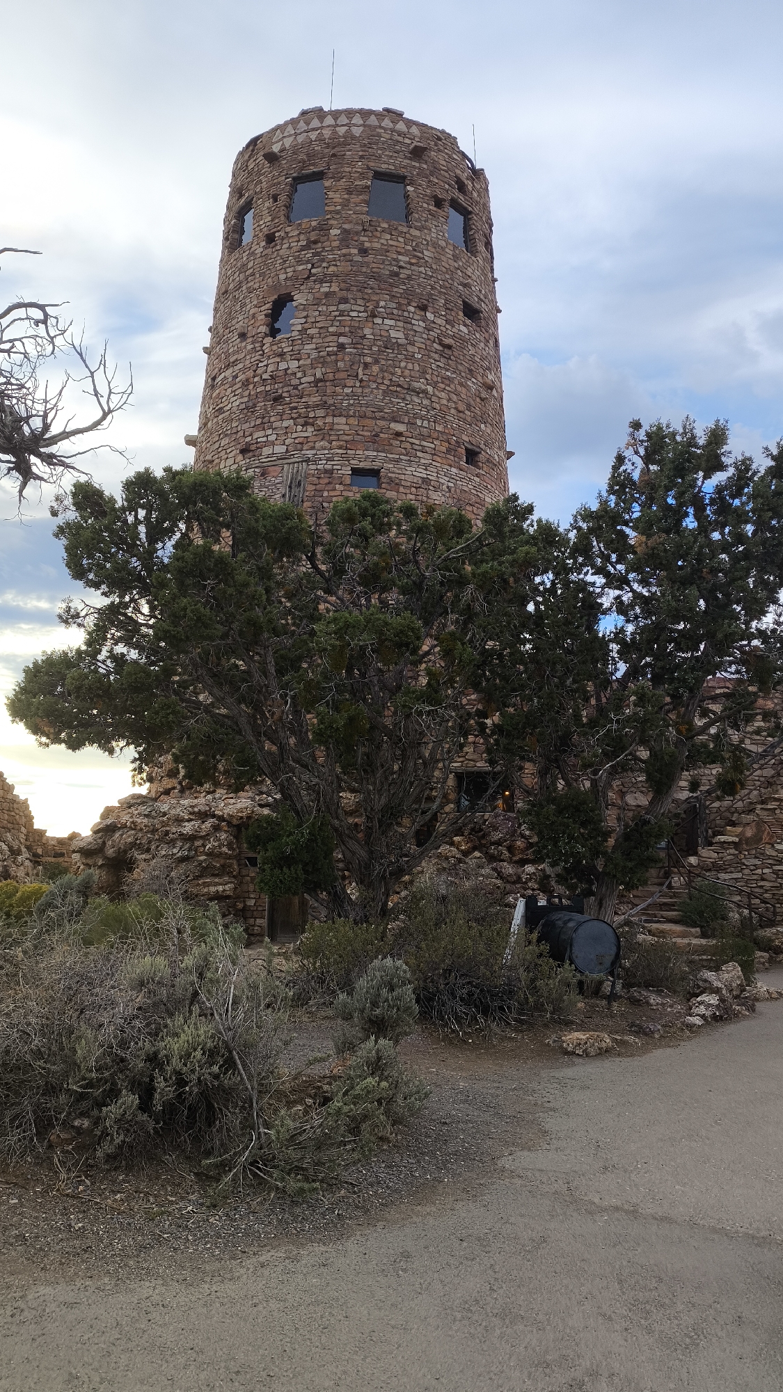The image shows a stone tower with a conical roof and multiple windows, set against a cloudy sky. The tower is surrounded by rocky terrain and sparse vegetation, including a large tree in the foreground. A pathway is visible in the lower right corner. The architecture of the tower suggests it is a historic structure, likely part of a national park or historical site. The natural elements and the presence of the tower are indicative of a location within the Grand Canyon, potentially Grand Canyon Village, Arizona, United States, a popular tourist destination known for its geological formations and historic architecture.
