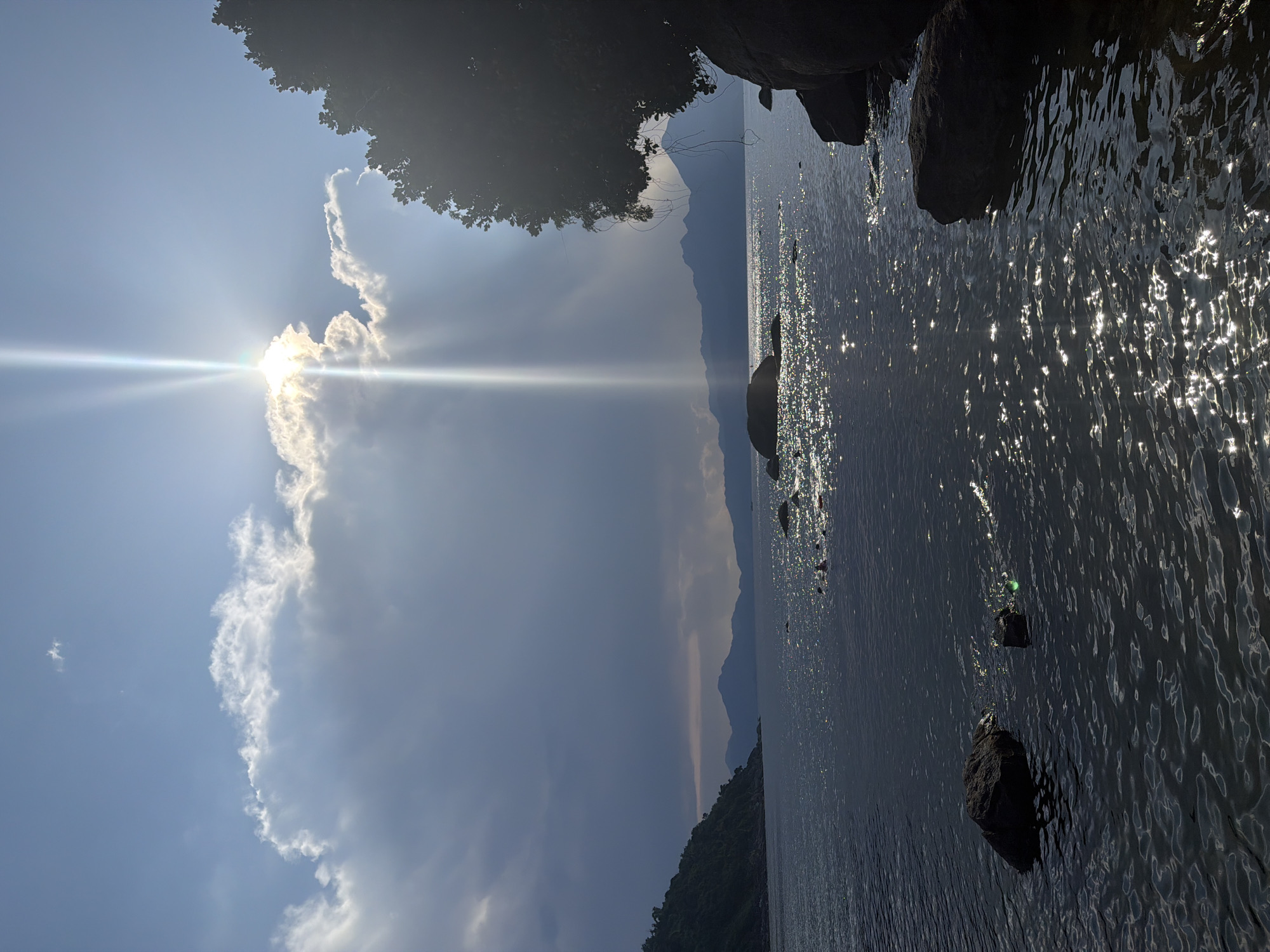 The image shows a landscape featuring a body of water, likely a bay or lake, with mountains visible in the distance. The foreground includes rocks along the shoreline and possible vegetation. The sun is obscured by clouds, creating a bright glare and sunbeams across the sky and reflecting off the water's surface.
Given the location being Đà Nẵng, Vietnam, it is likely this image depicts the Sơn Trà Peninsula, looking west across Đà Nẵng Bay towards the Bà Nà Hills. The rocks in the foreground would be the rocky coastline of the Sơn Trà Peninsula.