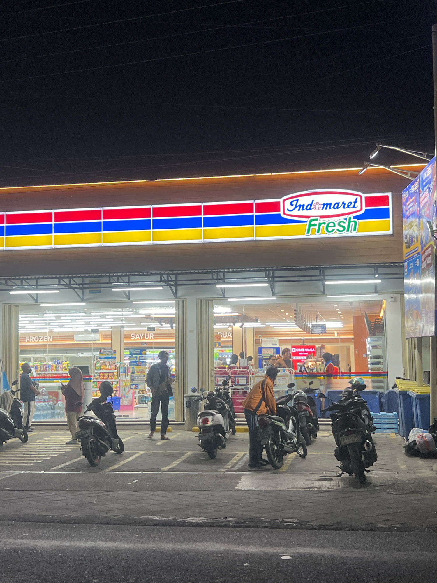This is a nighttime, eye-level, full-shot photograph of an Indomaret Fresh convenience store in Tlogoadi, Indonesia. The store's facade is brightly lit, with a prominent sign displaying "Indomaret Fresh" in red and green lettering against a white background, framed by red, blue, and yellow horizontal stripes. Inside the store, shelves stocked with various products are visible through the glass entrance.
In front of the store, several motorcycles are parked. Some individuals are standing near the motorcycles or entering/exiting the store. One person in an orange shirt is interacting with a motorcycle, while another person in a dark jacket stands further back. Two people in the foreground are standing near the entrance. The pavement in front of the store is marked with white lines, indicating parking spaces. Beyond the store, blue and yellow bins are stacked. The overall environment suggests a typical busy convenience store setting at night in a commercial area.