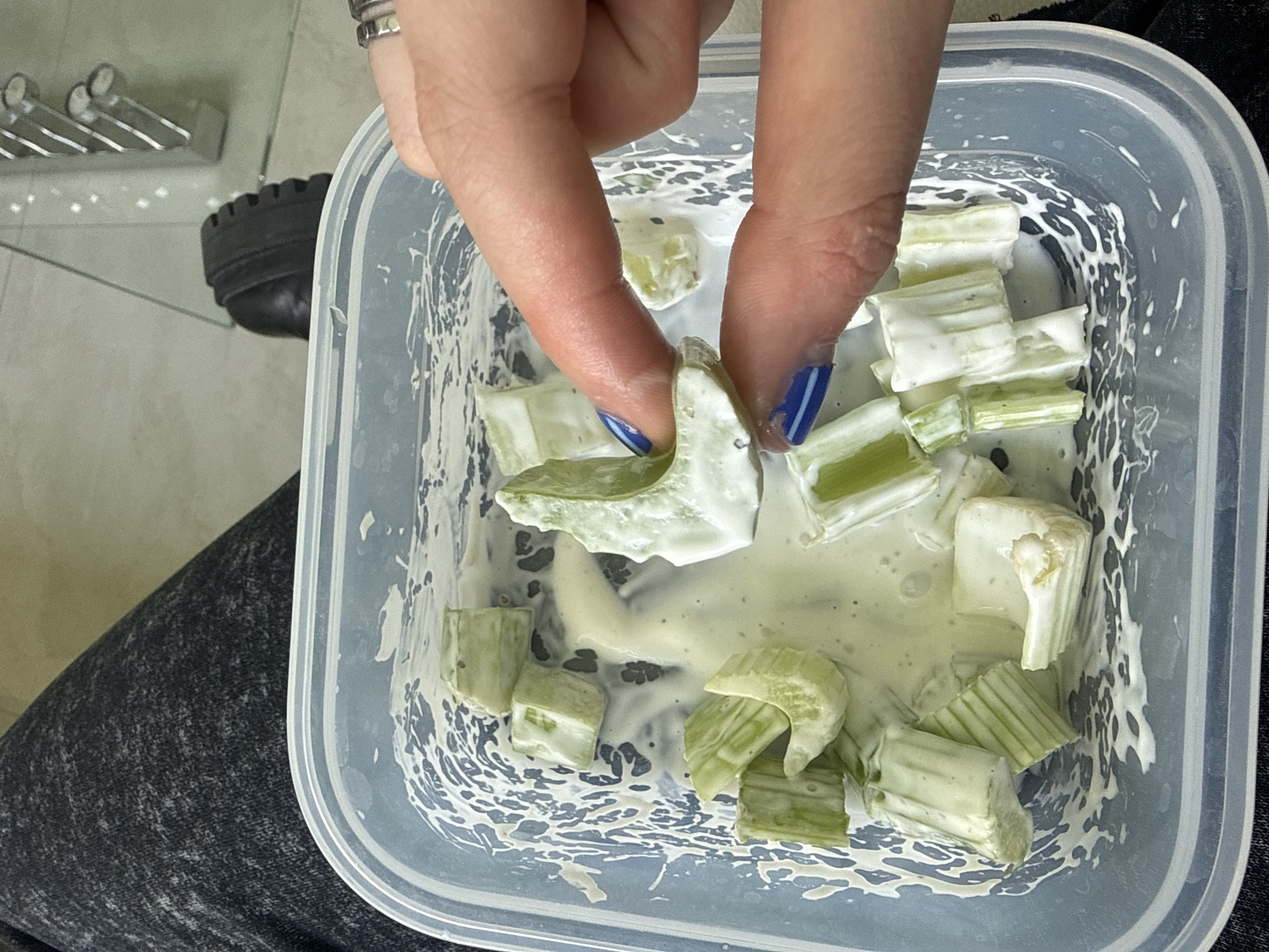 A person is holding a piece of celery covered in a white sauce over a container filled with more celery pieces and sauce. The person has blue nail polish with white stripes on their nails and rings on their fingers. The container is clear and rectangular, and the scene appears to be indoors. The background includes a dark grey fabric and part of a metal rack. There is no information to suggest this image was taken in Santa Clara Ocoyucan, Mexico.