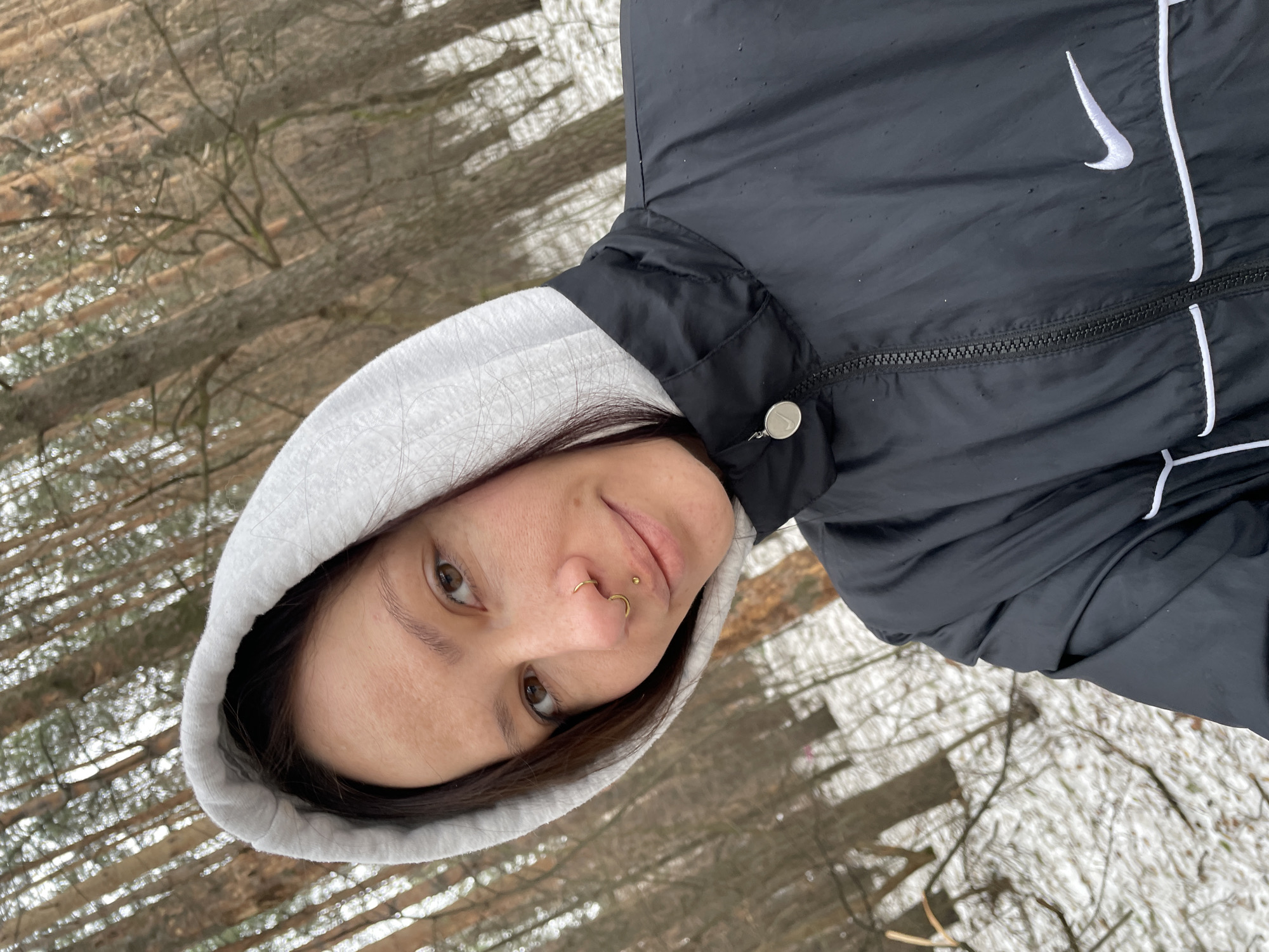 A woman is depicted from the chest up, appearing outdoors in a wooded area. She wears a gray hooded sweatshirt and a black Nike jacket. The background shows trees and a snowy ground. The photo was not taken in Izhevsk, Russia.