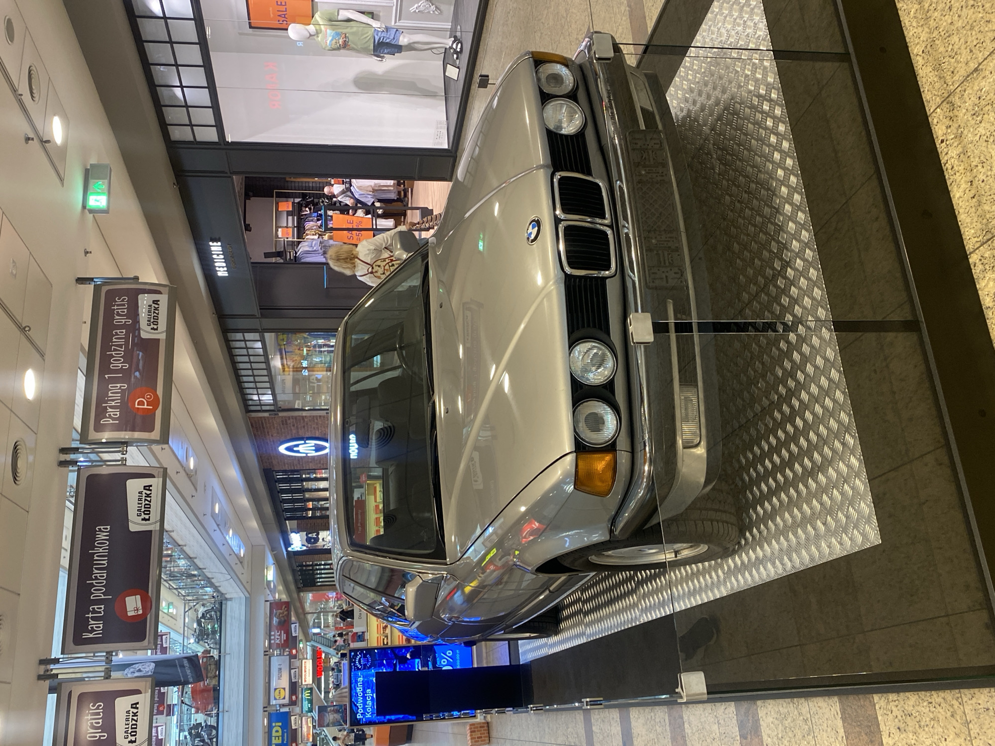 This media file shows an indoor scene, likely a shopping mall, with a vintage silver BMW on display. The car is positioned at an angle, with its front grille and headlights prominently visible. The setting appears to be a public space with shops and signage, suggesting a commercial environment.
Several signs are visible, indicating that the location is "GALERIA ŁÓDZKA" in Łódź, Poland. Some signs advertise "Parking 1 godzina gratis" (1 hour free parking) and "Karta podarunkowa" (gift card), suggesting these are amenities or services offered at the mall. Another sign reads "MEDICINE", which is likely a store name. There are also signs with prices like "SALE" and "%".
The lighting is artificial and appears to be typical for an indoor commercial space. There are hints of people in the background, possibly shoppers or mall staff, but they are not the primary focus. The overall impression is a car exhibition or promotional event within a shopping center, featuring a classic BMW. The image is oriented vertically.