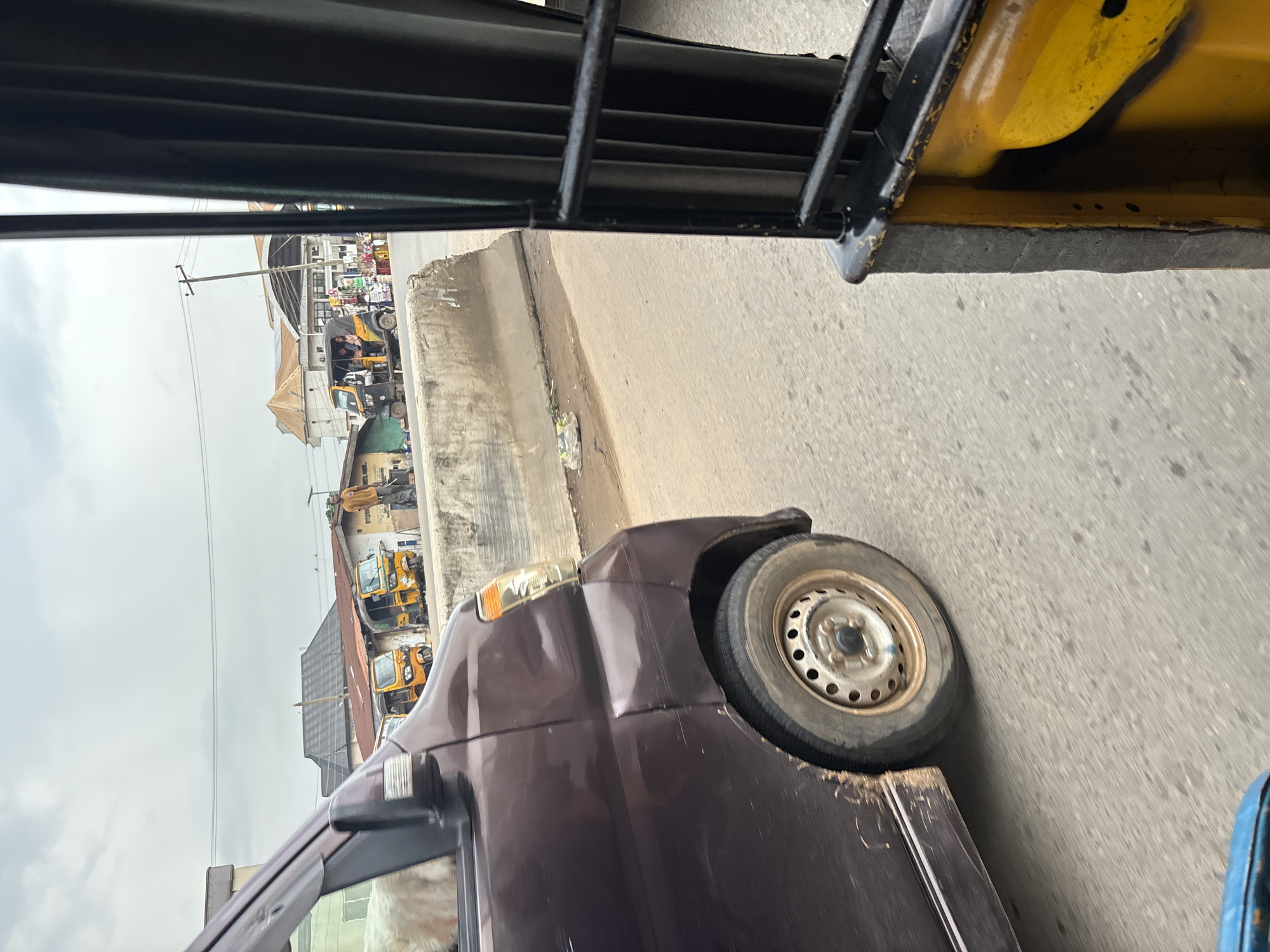 The image depicts a street scene in Onitsha, Nigeria, viewed from inside a vehicle. In the foreground, the side of a dark brown minivan is visible, showing its front wheel, mud splatter on the lower body, and a slightly damaged headlight. To the right, the interior of another vehicle, likely a yellow auto-rickshaw or a similar transport, is partially visible, featuring black bars and yellow interior paneling.
The street itself is paved with asphalt. A concrete barrier runs along the side of the road, separating it from a raised area where several yellow auto-rickshaws are parked or in motion. In the background, buildings with corrugated metal roofs are visible, along with a utility pole and electrical wires. Several people are present, including individuals standing near the auto-rickshaws and others riding in them. The overall atmosphere suggests a typical urban environment with active transportation and commerce. The sky is overcast, indicating potentially cloudy weather.