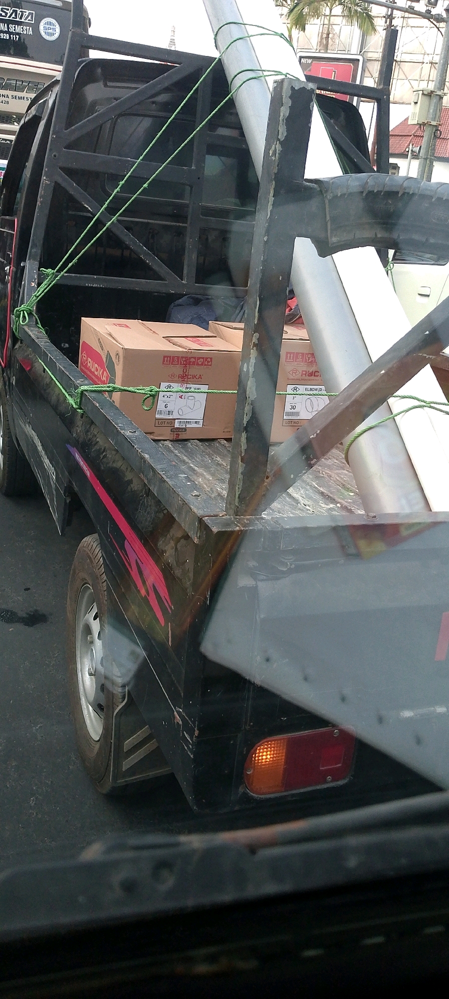 An outdoor medium shot captures a black pickup truck on a street in Bandung City, Indonesia. The truck bed is filled with large gray pipes and cardboard boxes. The pipes are secured to the truck with green ropes and metal framing. The truck's rear features a visible red and orange taillight and a reflective strip. The background includes buildings, trees, and a blue sky, suggesting daylight. The overall scene is a street view of a truck transporting materials.