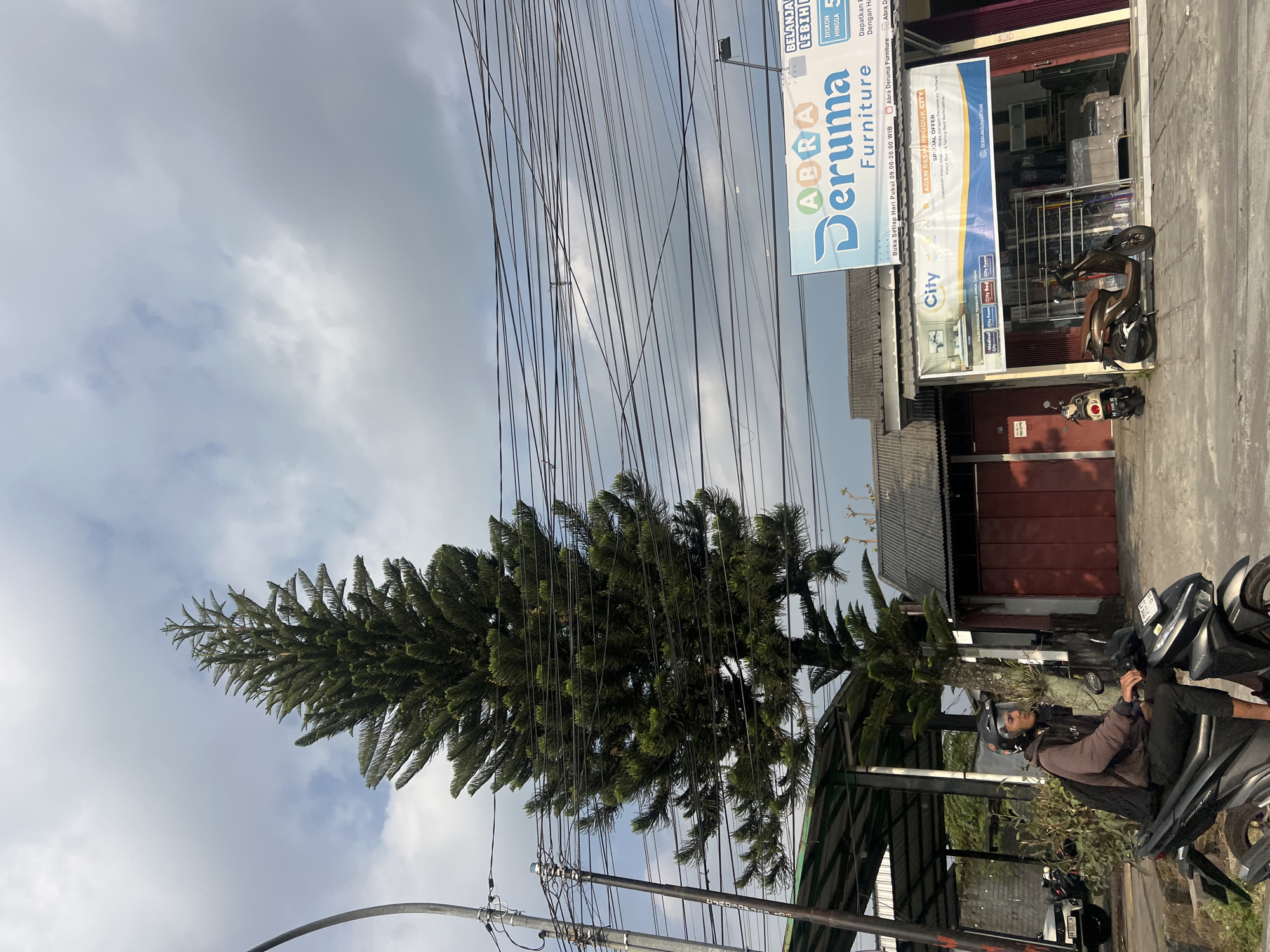A person wearing a dark hoodie and helmet rides a black scooter on a street in Ngaglik, Indonesia. The rider looks to their right. A tall, dark green pine tree stands to the left of the frame, its branches interwoven with a dense network of black electrical wires overhead. To the right of the tree, a building with a grey roof displays a sign that reads "ABRA Deruma Furniture." Another banner below it advertises "City" products. Two motorcycles are parked in front of the furniture store. The sky is overcast with grey clouds.