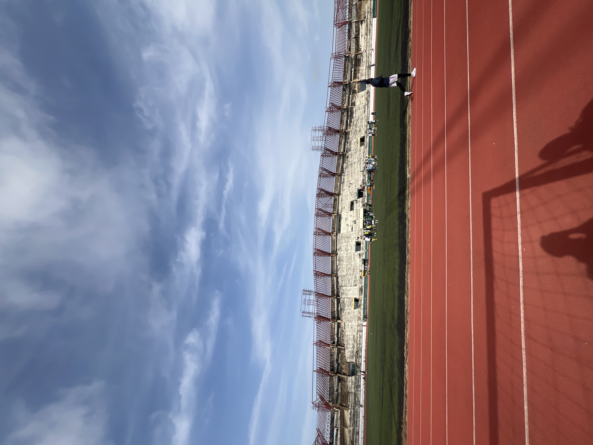 The image captures a wide view of the Ahmadu Bello Stadium in Kaduna, Nigeria, on a bright, partially cloudy day. The stadium features tiered seating, with sections appearing to be under renovation or disrepair, indicated by scaffolding and exposed concrete. The foreground is dominated by a red athletic track, with white lane markers clearly visible. A lone individual, dressed in athletic wear, is running on the track in the middle ground. Beyond the track is a green football pitch where a group of people, presumably footballers in training or a team, are gathered. The sky above is a vibrant blue, streaked with wispy white clouds. The overall scene suggests an active sports venue, possibly undergoing development.