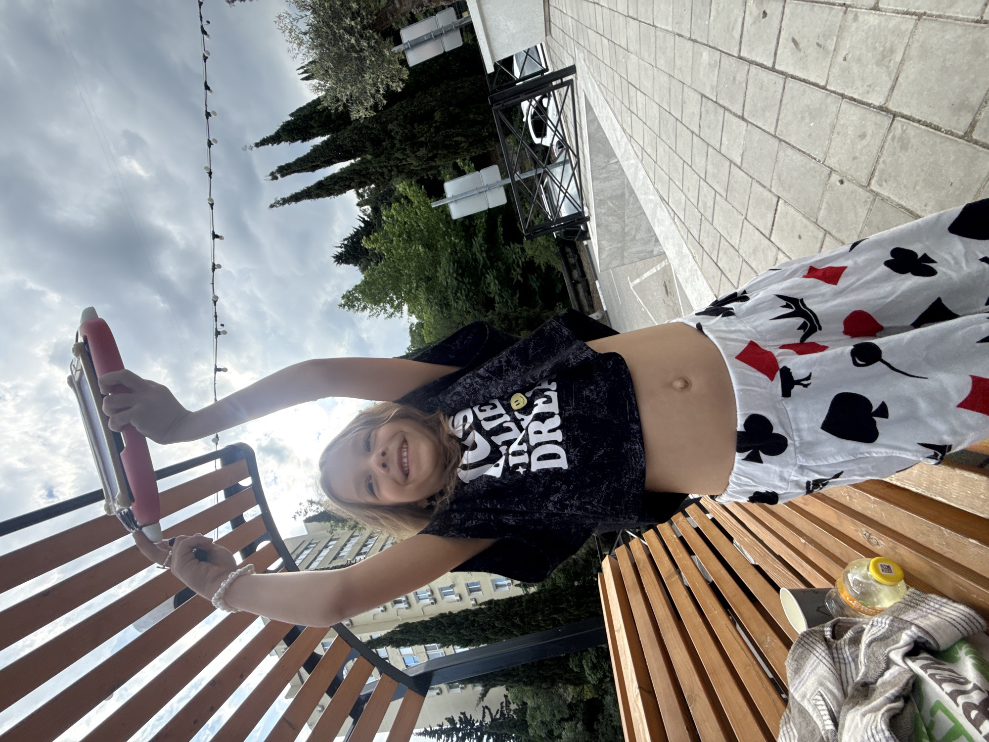 A young girl with blonde hair is pictured outdoors, holding a red and silver hair curler above her head. She is smiling and wearing a black crop top with white lettering and white pants with a black and red playing card pattern. The girl is seated on a wooden bench, with a blanket and a bottle of liquid on the bench next to her. In the background, there are trees, buildings, and a cloudy sky. The scene appears to be in a park or public area in an urban setting.