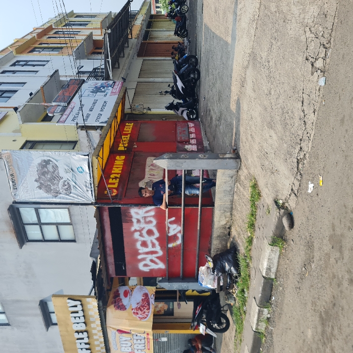 The scene takes place in front of a red and yellow storefront, labeled "Le2king Pegel Lele," in Dramaga, Indonesia (latitude: -6.574, longitude: 106.751). There is a young man in a blue shirt and jeans leaning on a metal railing. He appears to be looking off to the side. In the foreground, there is a black plastic bag and a few pieces of trash on the ground. There are a few motorcycles parked on the right side of the image. The building next to the storefront is pale yellow and has a sign that reads "Penjualan Barang, Kaos Merchandise, Sticker Photography," in Indonesian. The sidewalk is cracked and a bit uneven. The scene is a typical street corner in Indonesia.