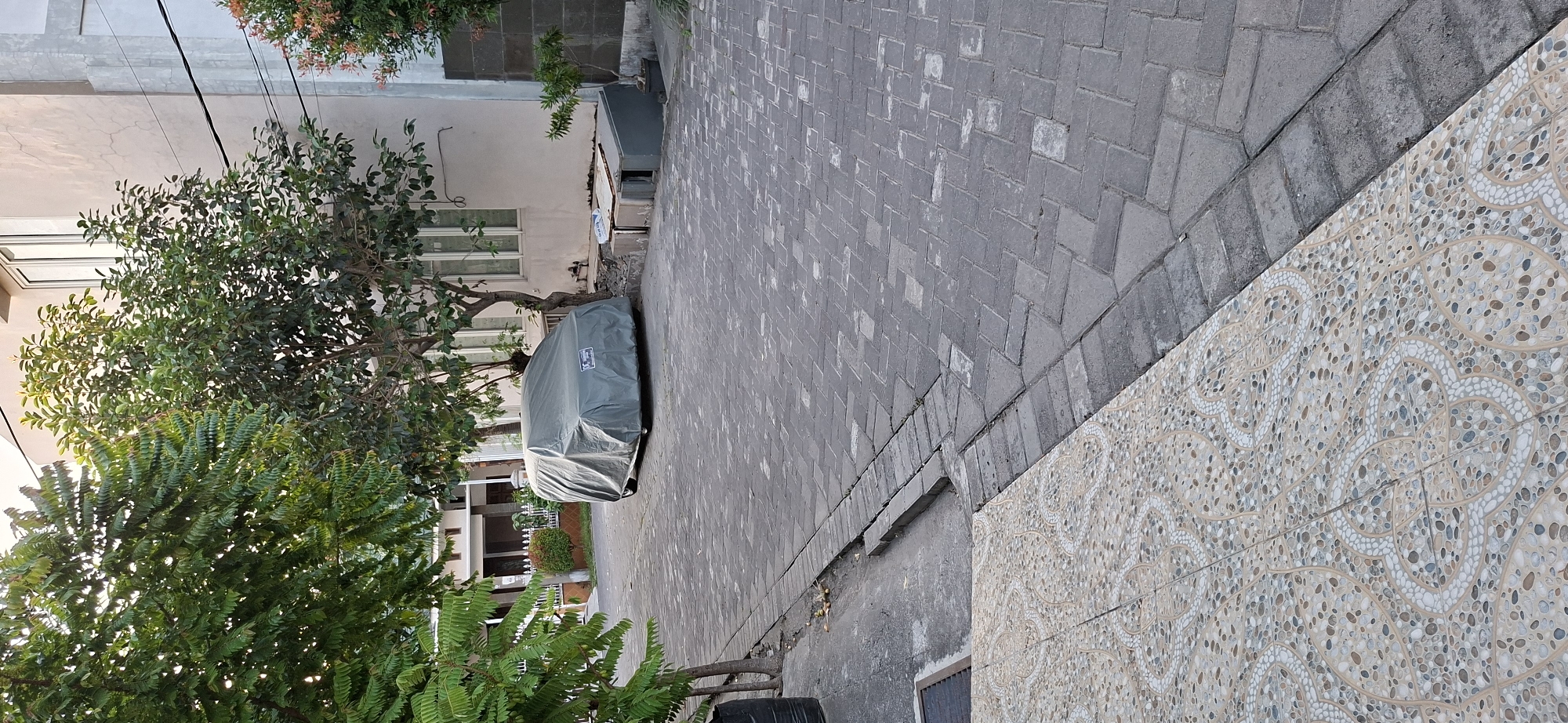 The image depicts a street scene in Surabaya, Indonesia. A car covered in a grey tarp is parked on a paved driveway. The driveway is made of interlocking grey bricks. To the left of the driveway, there are several trees with green foliage. In the background, a light-colored building with windows is visible. The weather appears to be overcast, as the light is diffused. The scene is quiet and still, with no people or animals present.
