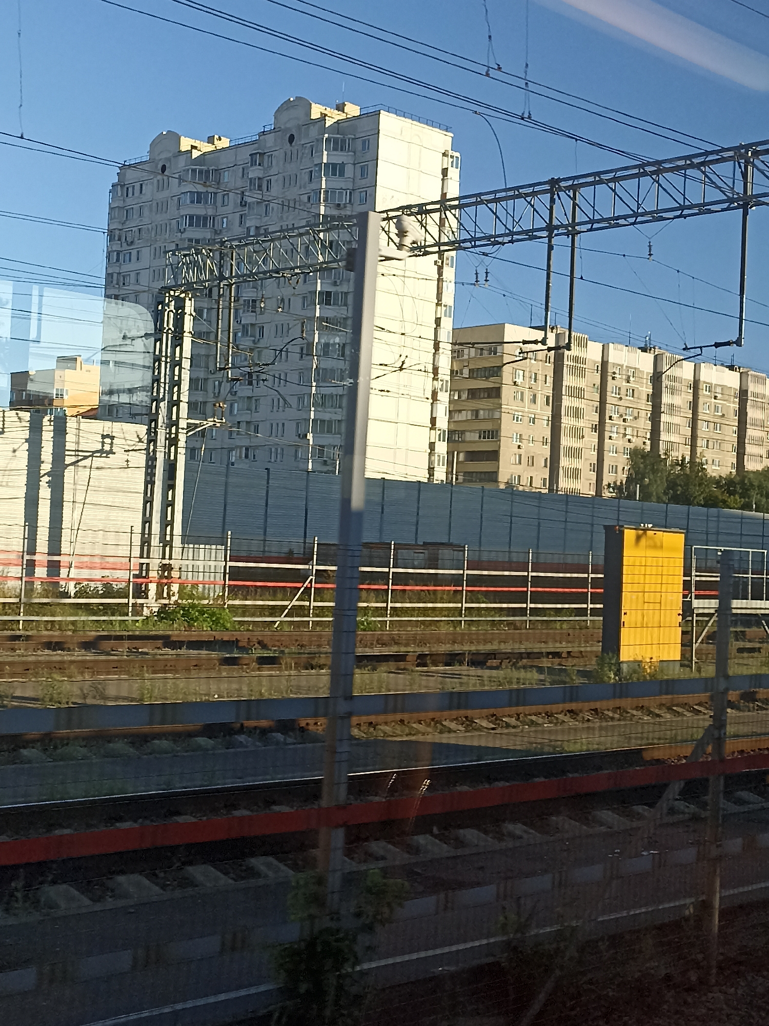 The image depicts a view from a train, likely in Podolsk, Russia, as indicated by the presence of a multi-story residential building and railway infrastructure. The sky is clear and blue. In the background, several apartment buildings with numerous windows are visible. In the middle ground, there is a blue noise barrier fence, and a yellow utility box. Railway tracks and overhead power lines dominate the foreground and midground, with some vegetation growing alongside the tracks. The overall scene suggests an urban environment with active transportation infrastructure.