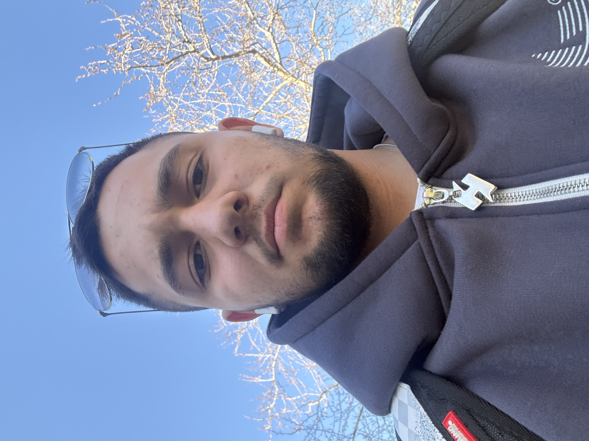 A man is standing outdoors on a sunny day. He is wearing a navy blue hooded sweatshirt, sunglasses on his head, and white wireless earbuds. Part of a tree with bare branches is visible in the background, along with a clear blue sky. The man appears to be taking a selfie or photo of himself. The location is unknown and cannot be determined from the image.