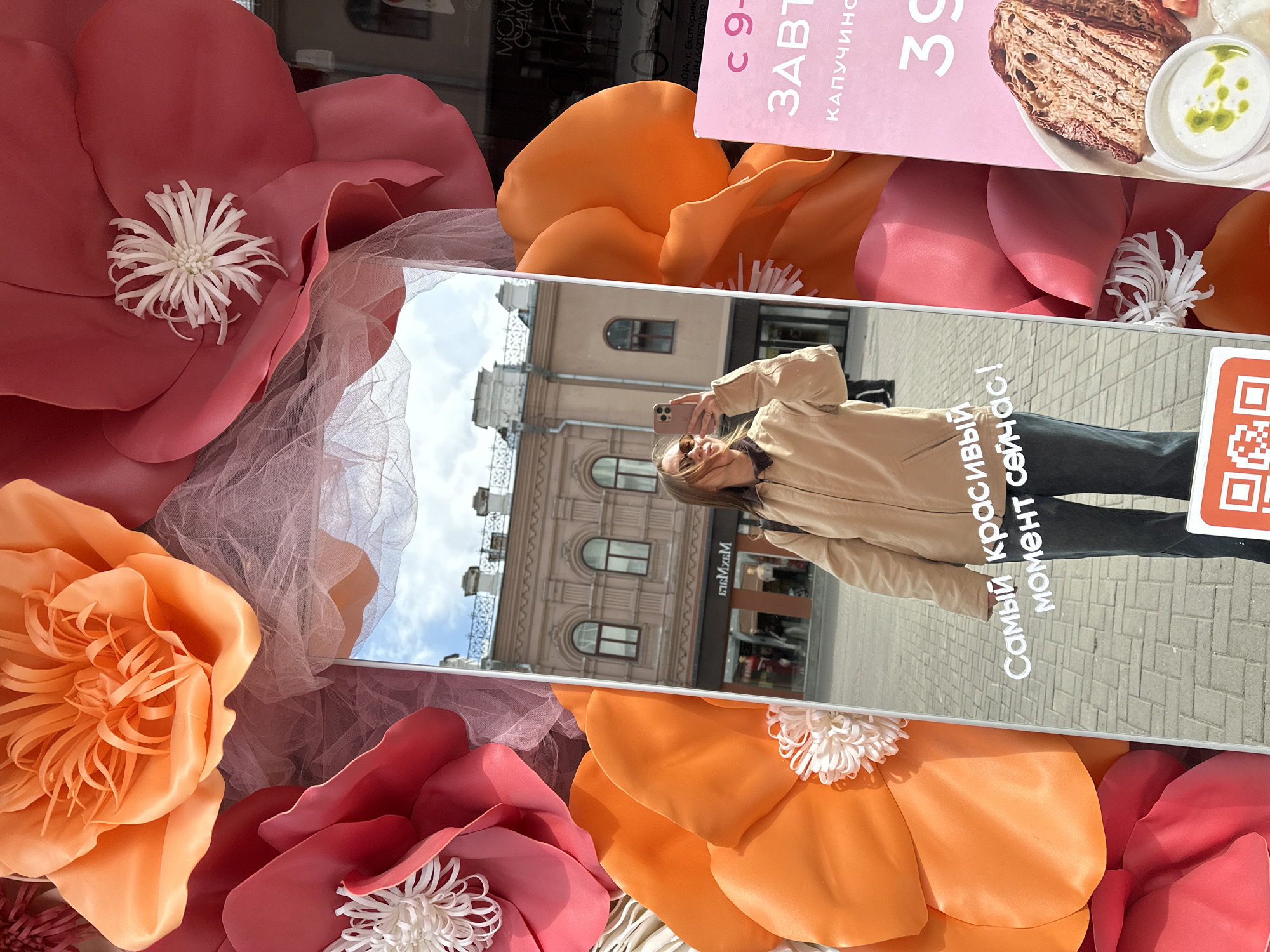 Here is a detailed description of the image:
**Scene:** The image captures a person taking a selfie in front of a mirror display decorated with large, colorful artificial flowers. The display appears to be set up on a city street.
**People:** A woman is visible in the reflection of the mirror, holding a smartphone and presumably taking a selfie. She is wearing sunglasses and a light-colored jacket.
**Location:** The scene is in Yekaterinburg, Russia. This is confirmed by the Russian text "г. Екатеринбург" in the top right corner, visible near a breakfast menu and a price of 39. The reflection also shows a building with a "Max Mara" sign, which can assist in identifying the specific street in Yekaterinburg.
**Objects:** Key objects include:
* The mirror display itself, framed by large artificial flowers in shades of pink and orange.
* The woman's smartphone.
* Buildings reflected in the mirror.
* A breakfast menu with a picture of bread and cream cheese.
**Environment:** The weather appears to be clear and sunny, as seen in the sky reflected in the mirror. The city environment includes buildings with architectural details. The surface that the mirror is on consists of grey brick.
