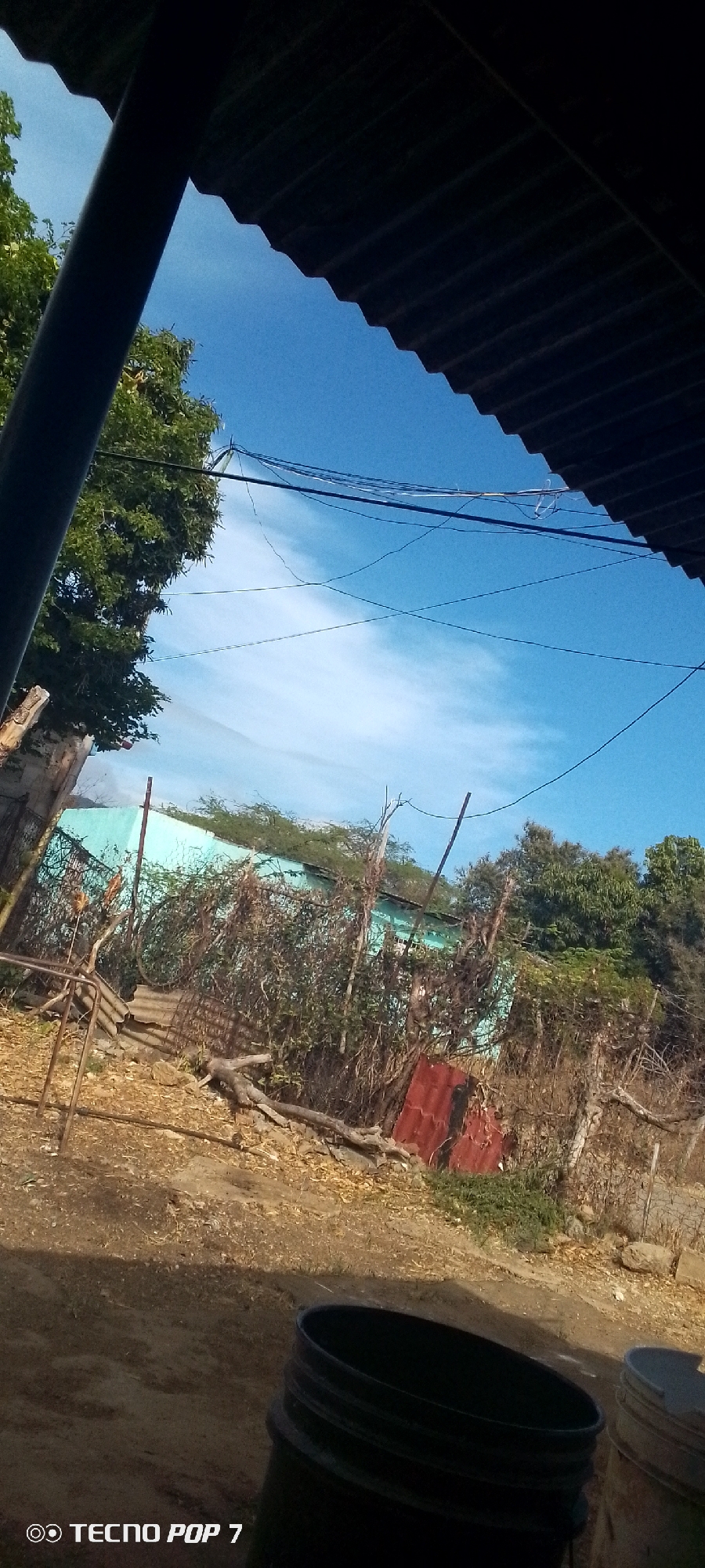 The image depicts an outdoor scene in Altagracia, Venezuela. The view is partially obstructed by a corrugated metal roof and a support beam. A blue sky with light clouds is visible above. The foreground shows a dirt ground with several black plastic containers. A fence line with vegetation is present, with a turquoise-painted building in the background. Discarded metal and wood debris are scattered in the midground. Power lines are strung across the sky. A "TECNO POP 7" watermark is visible in the lower left corner of the frame.