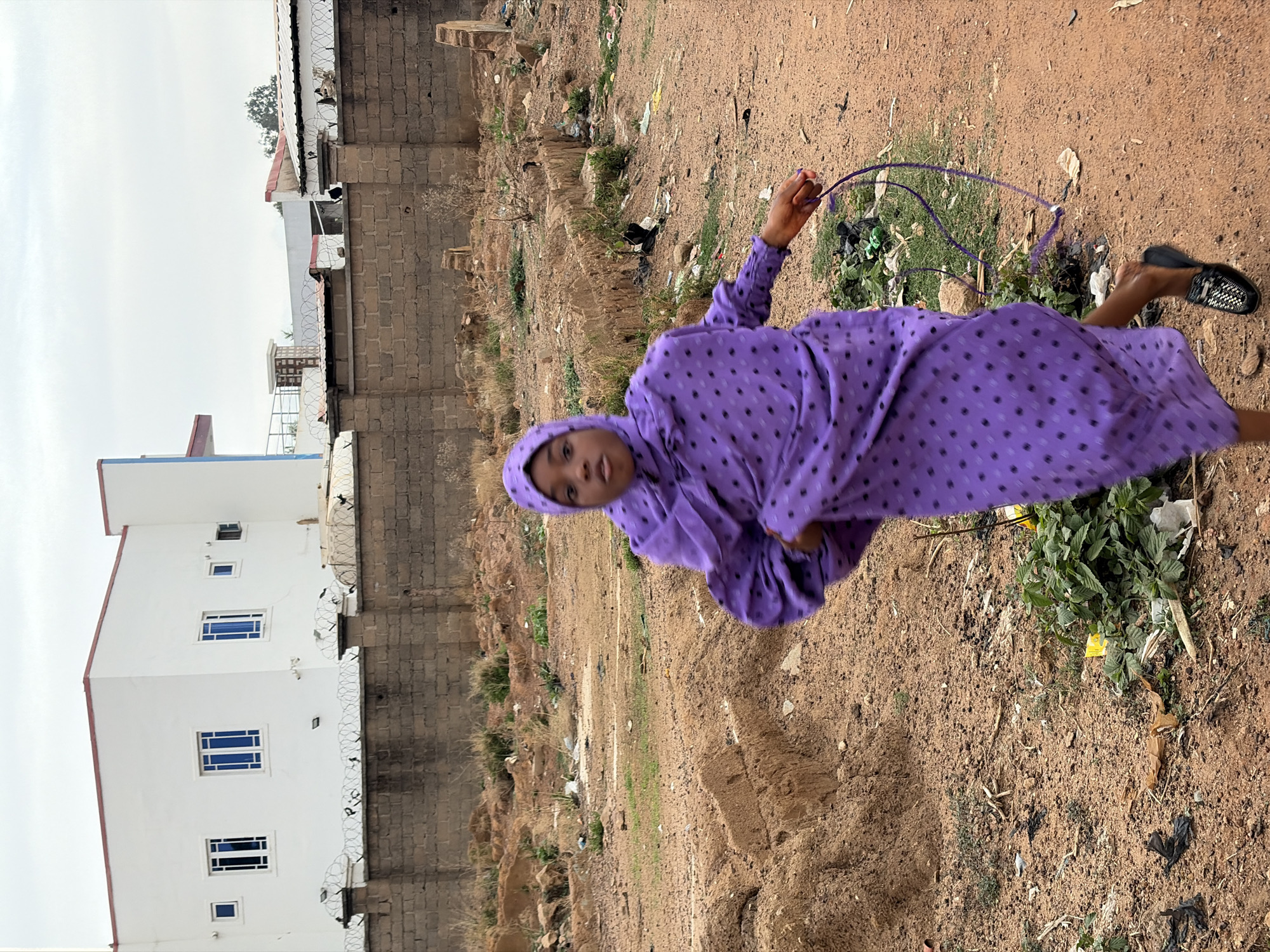A person wearing a purple polka dot dress and head covering stands in a sandy, trash-strewn area. The person is holding a purple string. In the background, there is a white building with blue-tinted windows, and a grey brick wall topped with barbed wire. The setting appears to be an undeveloped area, possibly in Barakin Lerawi, Nigeria.