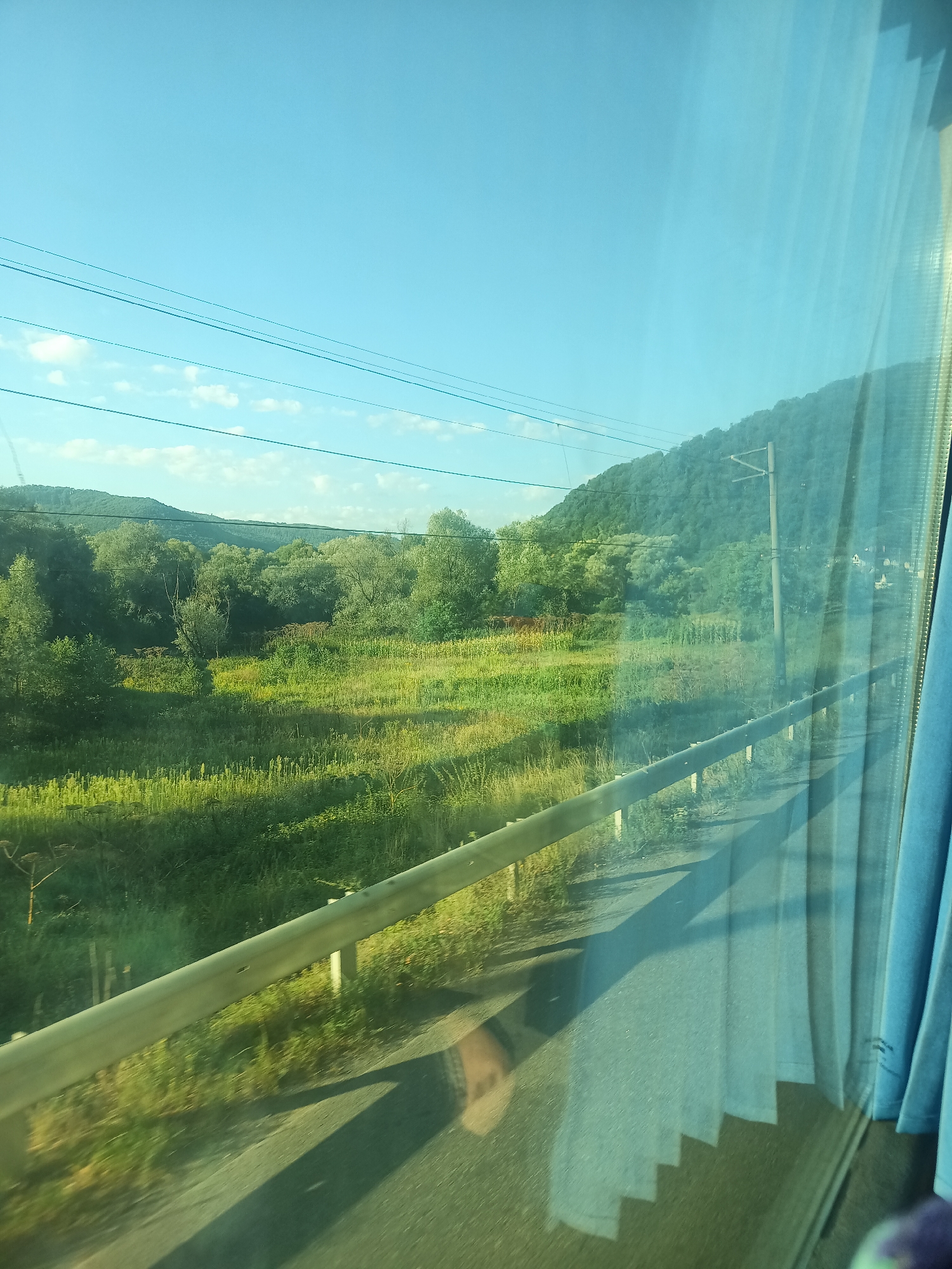 The image depicts a view from a moving vehicle, likely a train, through a window. The scene outside is a lush green landscape with rolling hills covered in trees. In the foreground, a metal guardrail runs parallel to the road, which is visible as asphalt. Power lines stretch across the bright blue sky. The window reflects the blue curtains of the vehicle and hints of a person's head. The image was taken in Perechyn, Ukraine, and captures a sunny day with scattered clouds.