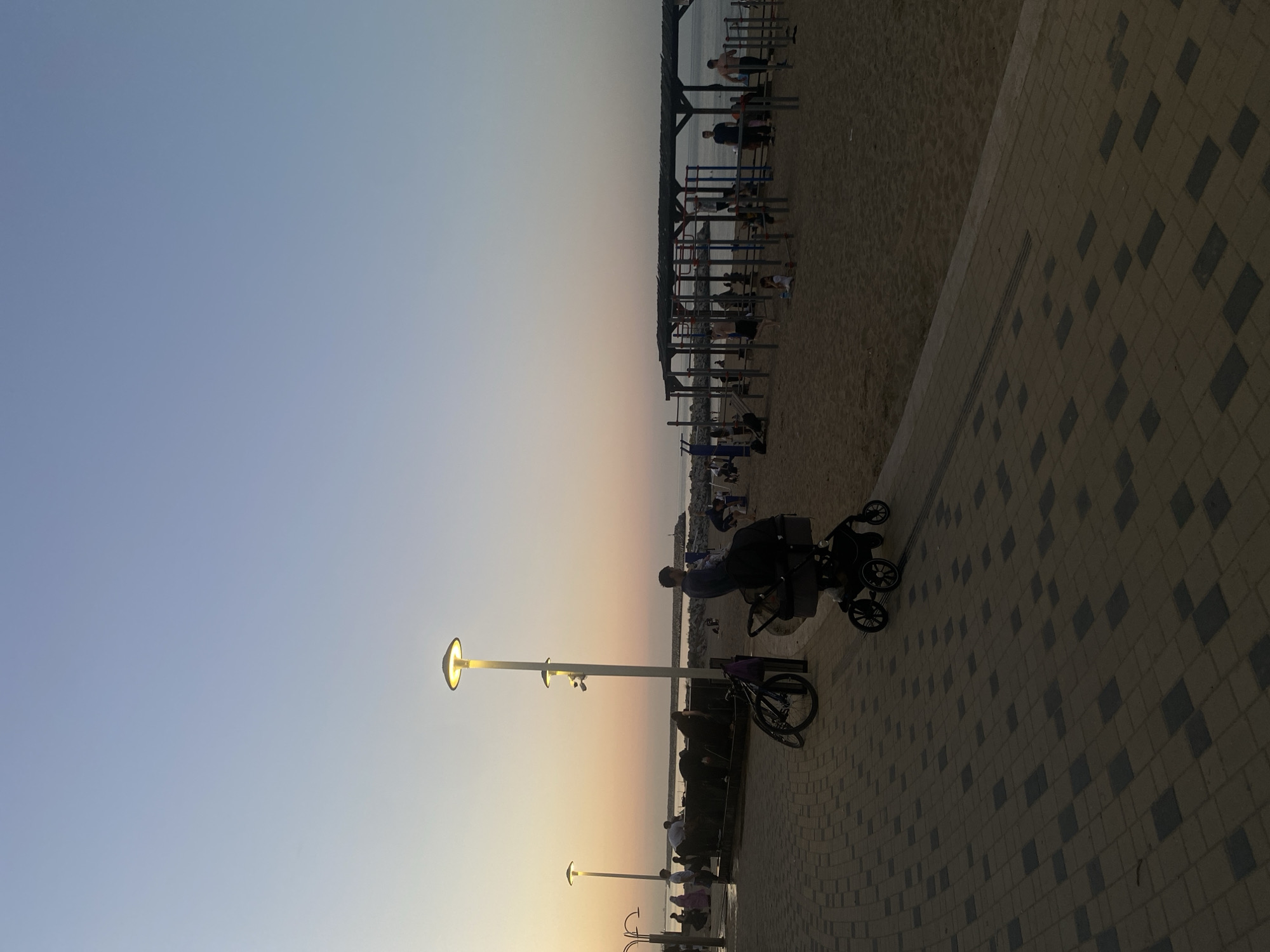 This is a photograph taken at dusk on a paved promenade adjacent to a sandy beach. The sky is a gradient of light blue, transitioning to a soft orange near the horizon where the sun is setting. In the foreground, a person is pushing a black stroller along the promenade. Further down the promenade, a bicycle is parked next to a lamppost. A group of people are visible near a series of outdoor showerheads, some appearing to be washing their hands or faces. In the background, on the beach, is an outdoor gym with various exercise equipment, and several individuals are engaged in physical activity. The sea is visible beyond the beach, with a rocky breakwater extending into it. The overall atmosphere is peaceful, with people enjoying the beach and promenade in the evening light. This scene is identified as being in Makhachkala, Russia.