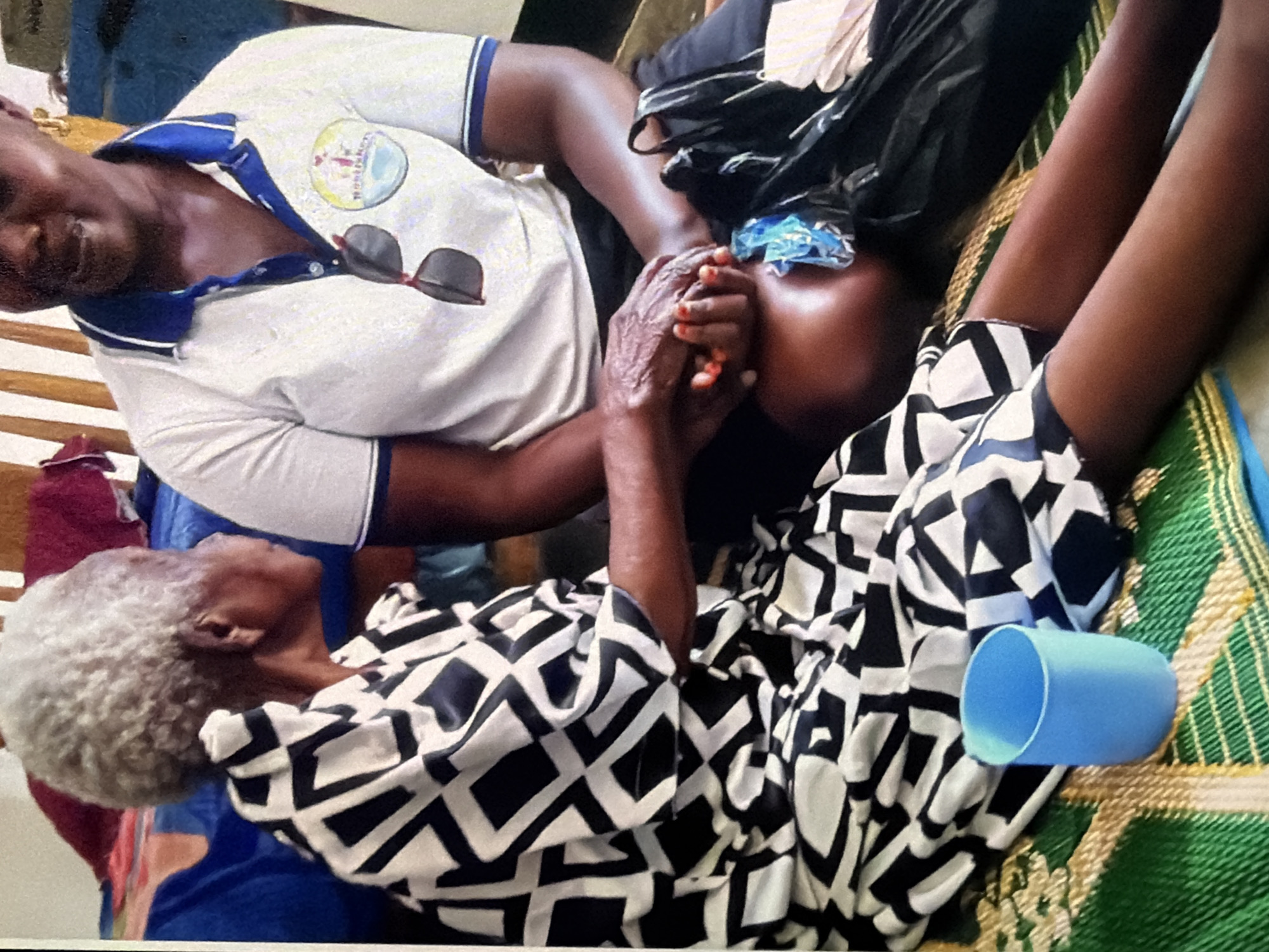 Two individuals, one elderly with grey hair wearing a black and white patterned dress, and another younger person wearing a white polo shirt with blue trim and sunglasses perched on the collar, are seated on a mat on the floor. Their hands are clasped together, suggesting an interaction of comfort or connection. A light blue cup sits on the mat in front of the elderly individual. Behind them, a bed with colorful bedding is visible, along with wooden slats of furniture. The environment appears to be indoors, possibly a home or community space.