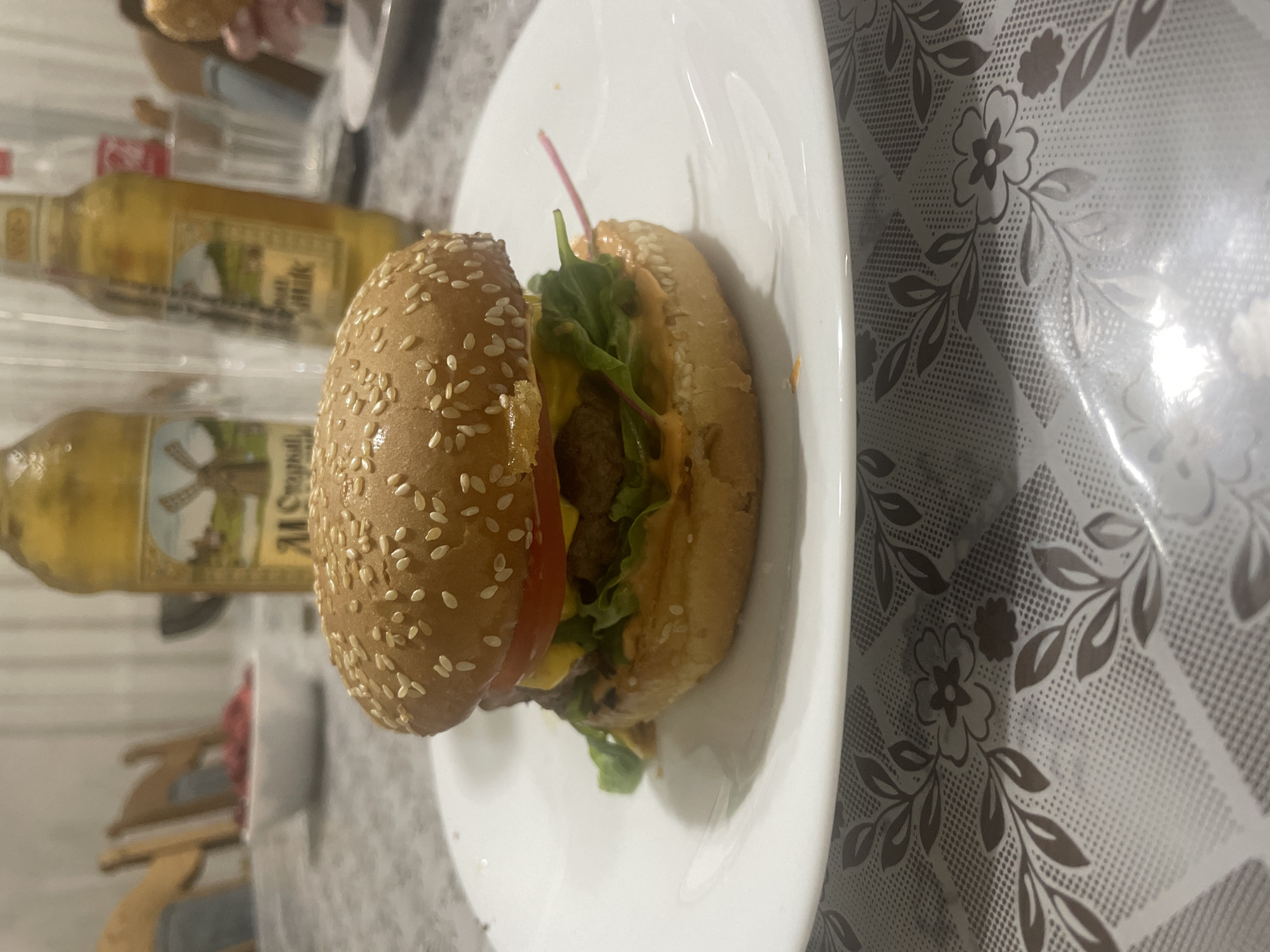 A close-up, eye-level, medium shot captures a hamburger on a white plate, placed on a table covered with a floral patterned tablecloth. The hamburger, the primary subject, is in focus and centered on the plate. It consists of a sesame seed bun, lettuce, tomato, and a patty, with a creamy orange sauce visible on the bottom bun. In the background, out of focus, are two glass bottles of beer with labels featuring windmills, a white bowl containing what appears to be chopped meat, and glimpses of wooden chairs and a table. The overall lighting is dim, suggesting an indoor setting, possibly a home or casual dining establishment. The scene is devoid of any identifiable city or location markers.