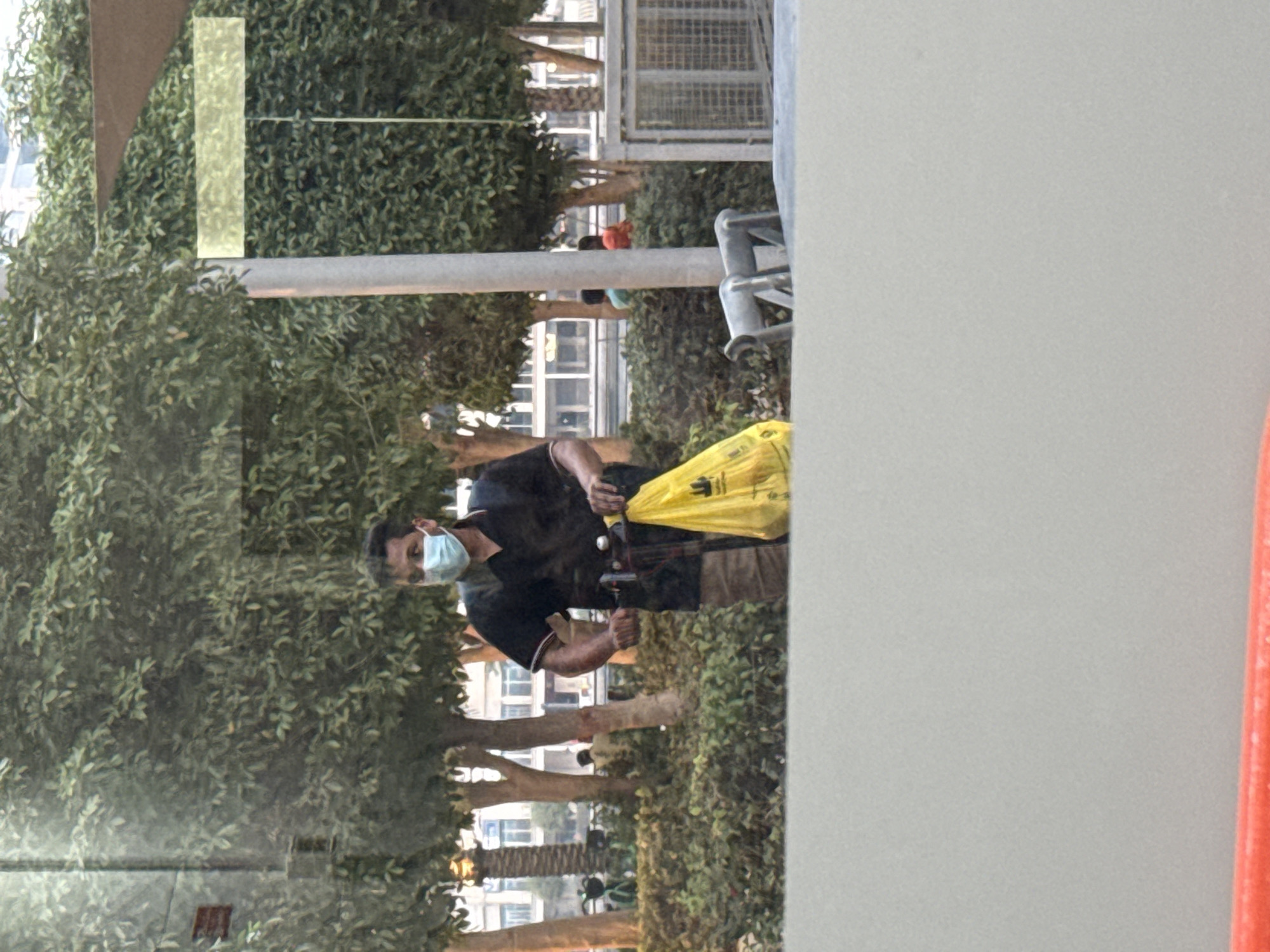 A man wearing a light blue face mask, black polo shirt, and patterned shorts is riding a scooter. He is holding a yellow shopping bag in his right hand. The man is in an outdoor urban setting, possibly in Dubai, United Arab Emirates, with trees and buildings visible in the background. The scene appears to be a public space with a paved walkway or area where people can ride scooters. A light-colored wall or barrier is in the foreground, obscuring the lower part of the image. The overall lighting suggests it could be late afternoon or early evening.