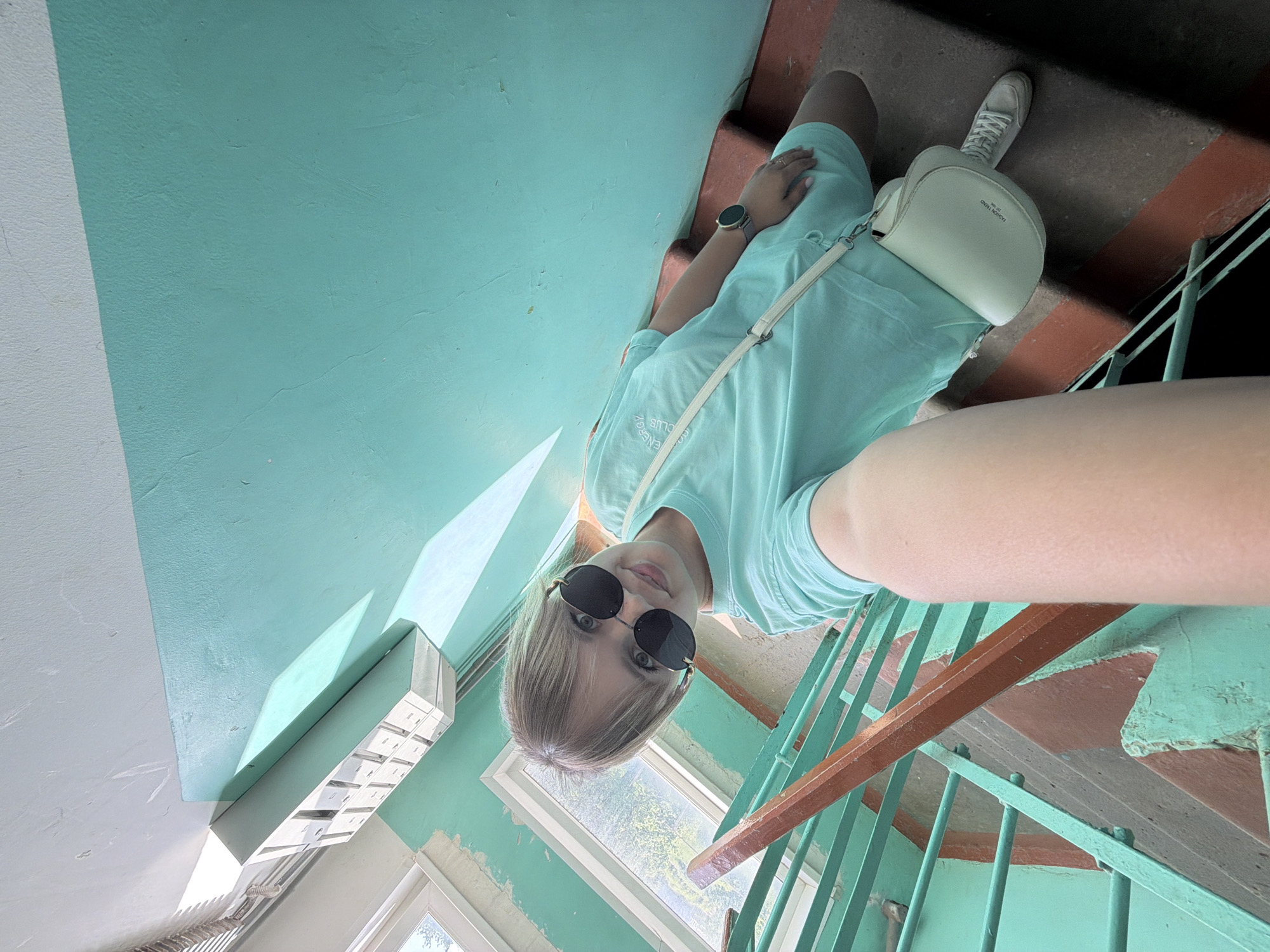 The image is a low-angle selfie taken on a stairwell. A young woman is centered in the frame. She has short blonde hair, wears round sunglasses, and is dressed in a light blue t-shirt and shorts. She is also wearing a watch on her left wrist and has a small white purse slung across her body. The background consists of a blue wall, windows with green frames, and stairs with brown railings. The location is in Chaikovsky, Russia.