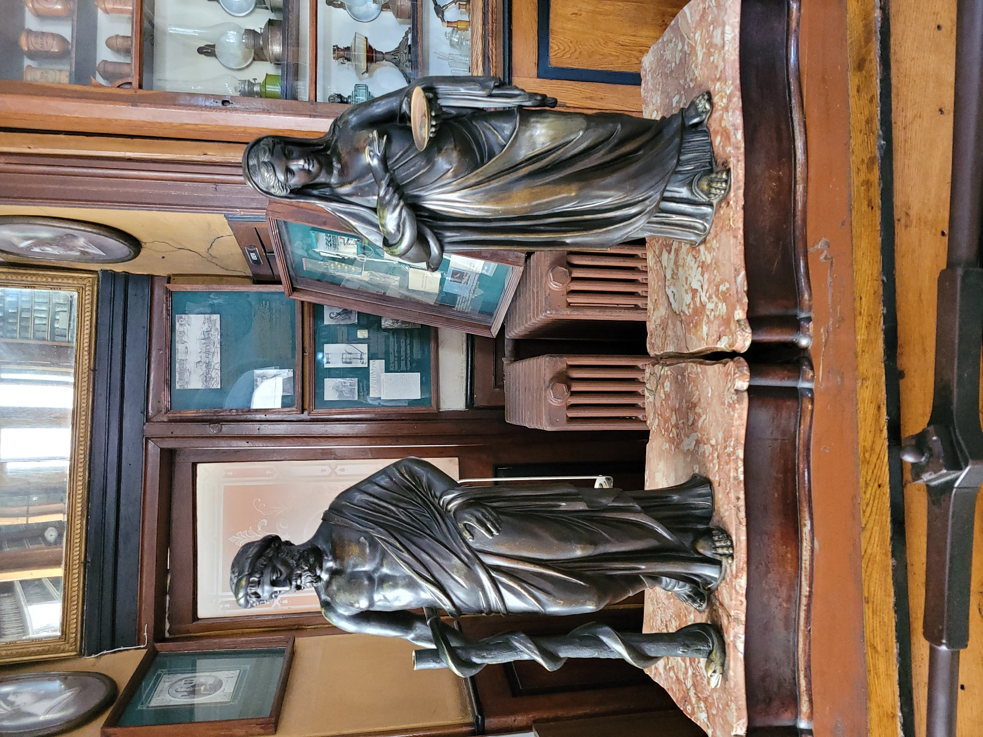 The image shows two bronze statues, a male and a female, on display in what appears to be a museum or antique shop. The male statue stands to the left, depicting a muscular man with a beard, draped in a toga, and holding a staff with a snake coiled around it. The female statue stands to the right, also draped in a toga, holding a small plate in one hand and a snake in the other. Both statues are placed on marble pedestals atop wooden cabinets. In the background, there are wooden cabinets filled with glass jars and scientific instruments, possibly indicating a historical pharmacy or medical display. Framed pictures and mirrors hang on the walls. The overall setting suggests an indoor environment with antique furnishings and historical artifacts. The location is identified as Unknown, Unknown.