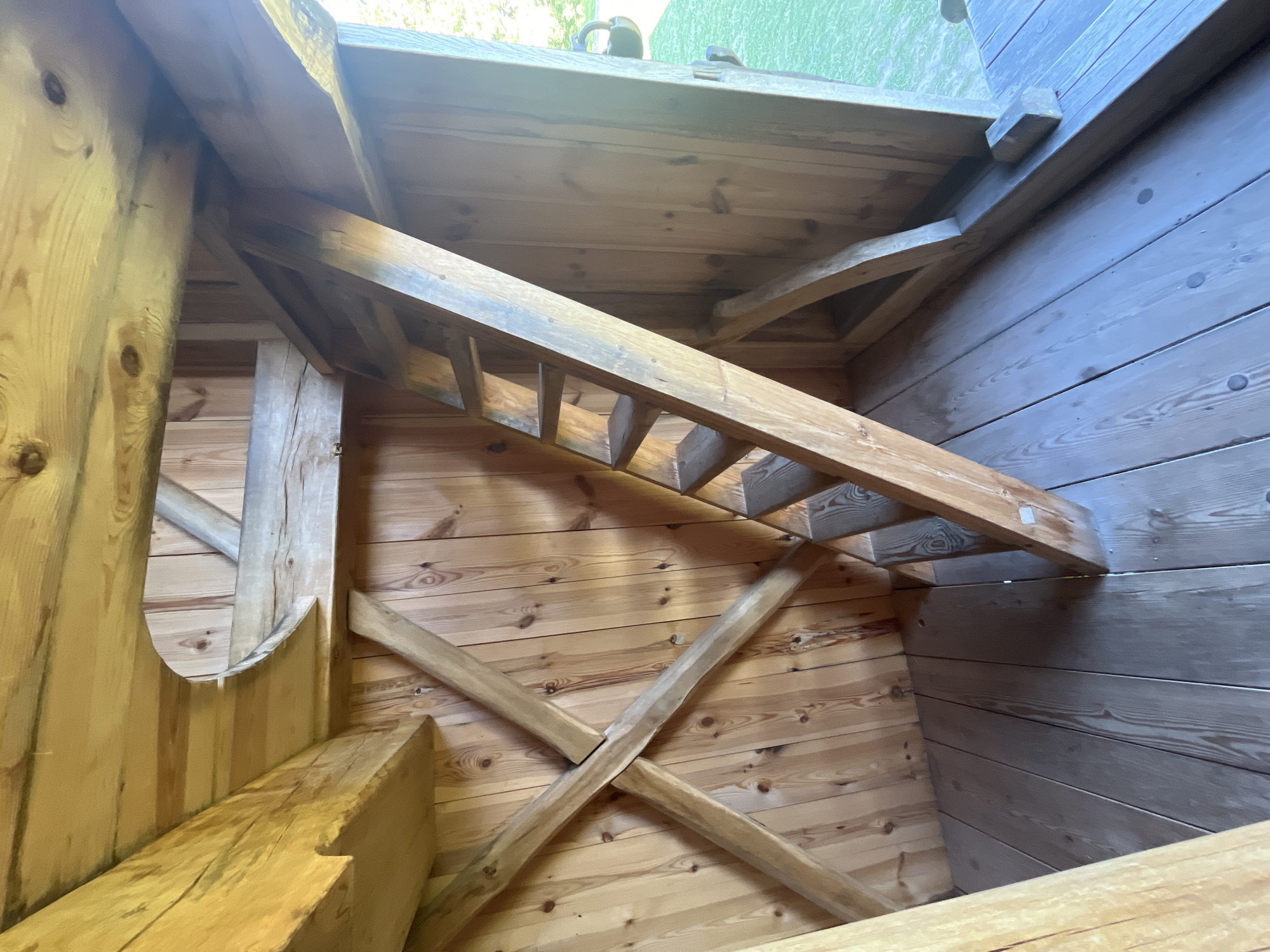 The image shows the interior of a wooden structure, likely a playhouse or a small shed, with visible exposed wooden beams and plank walls. A wooden ladder leans against the wall, leading up to an opening. The floor is also made of wooden planks. In the background, through an opening, a patch of green grass and some foliage can be seen, suggesting an outdoor setting. The lighting indicates it is daytime, with natural light entering the structure. There are no people or animals visible in the image. The architectural style appears rustic and handmade.