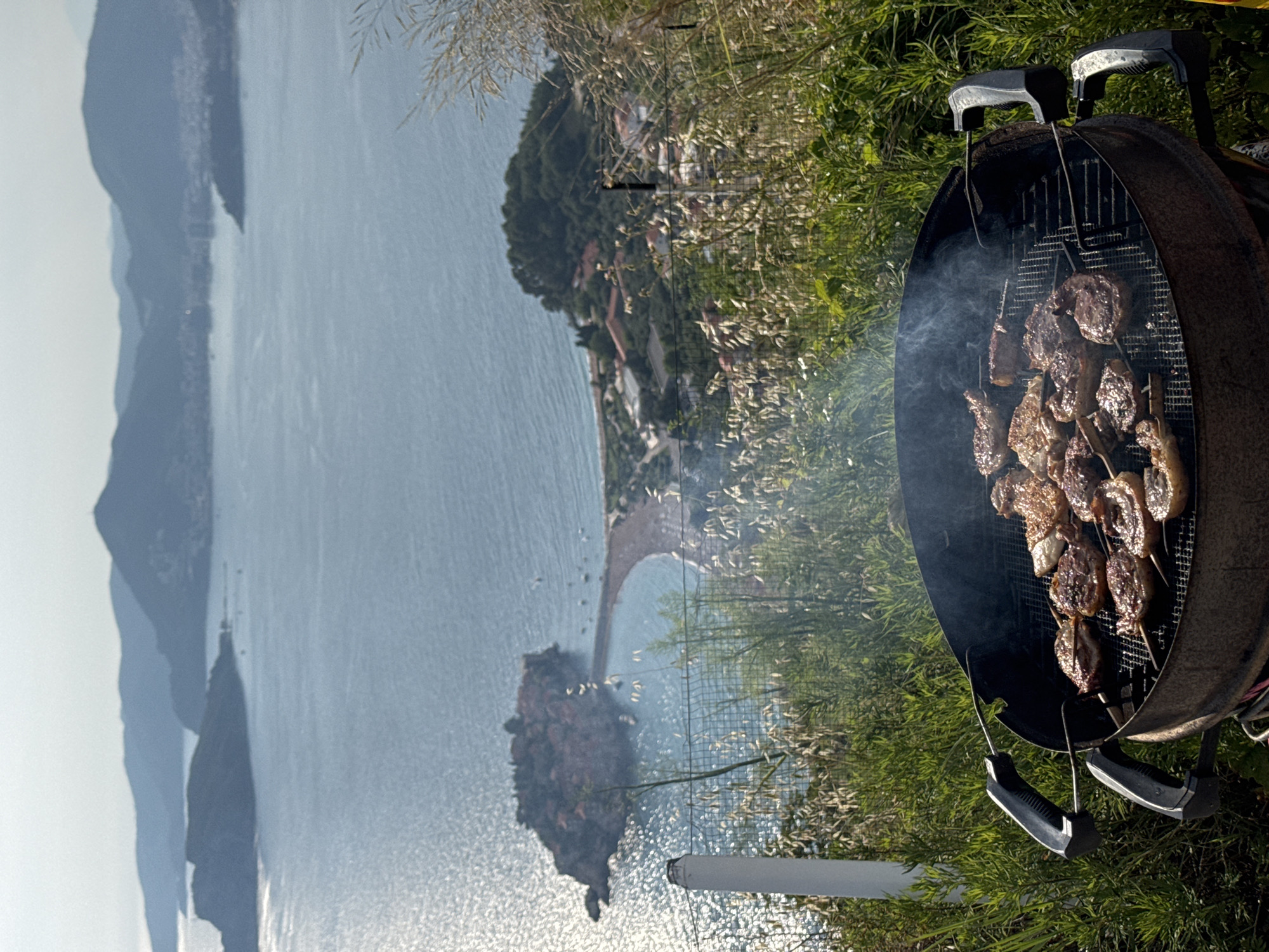 In Sveti Stefan, Montenegro, a barbecue is underway on a grill, positioned at a high vantage point overlooking a scenic coastal landscape. The grill, a circular black metal Weber style, contains numerous skewers laden with meat, visibly cooking over coals. Smoke rises from the grill, indicating the cooking process. The foreground is dominated by the grill and surrounding lush green vegetation and tall dry grasses. In the background, the azure Adriatic Sea stretches out, dotted with distant islands and the characteristic red-roofed buildings of Sveti Stefan island, connected to the mainland by a narrow causeway. The overall scene captures a leisurely outdoor meal with a breathtaking view.