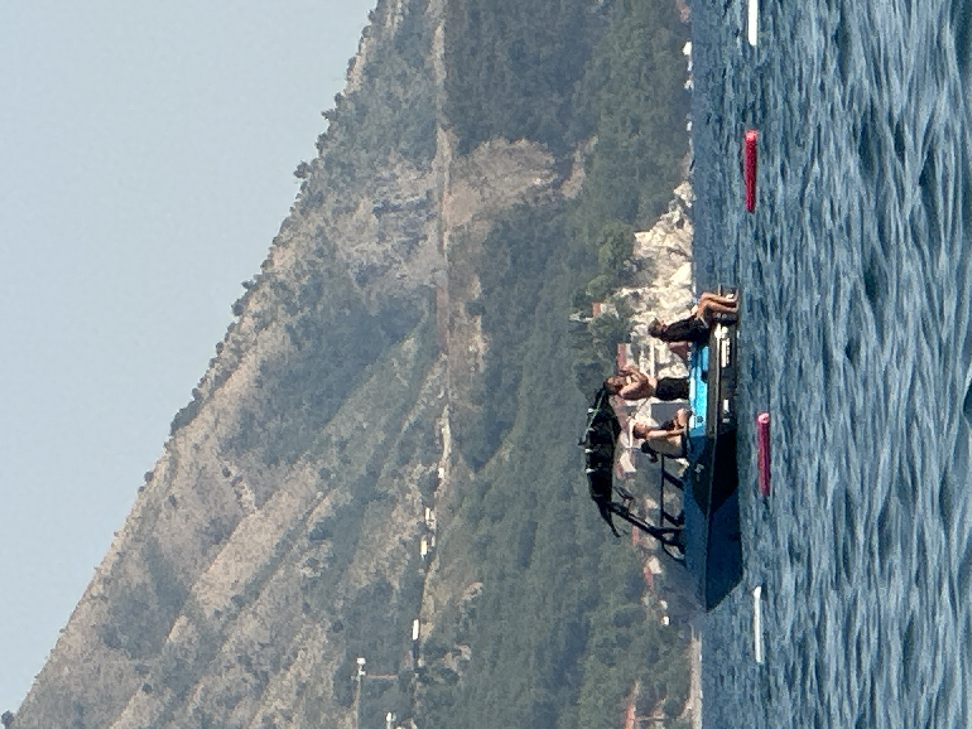 A blue and black motorboat with a canopy is positioned on choppy blue water in front of a steep, rocky, tree-covered mountain. Four individuals are visible on the boat. Two are standing in the rear of the boat, one is sitting on the edge of the boat, and another is sitting inside the boat. Three inflatable rafts, one pink and two white, are floating on the water in the foreground. In the middle ground, houses are visible on a rocky outcrop, and a tall construction crane is seen on the mountainside in the distance. The sky is clear and pale blue. The scene appears to be the coast near Boreti, Montenegro, given the characteristic mountainous landscape.