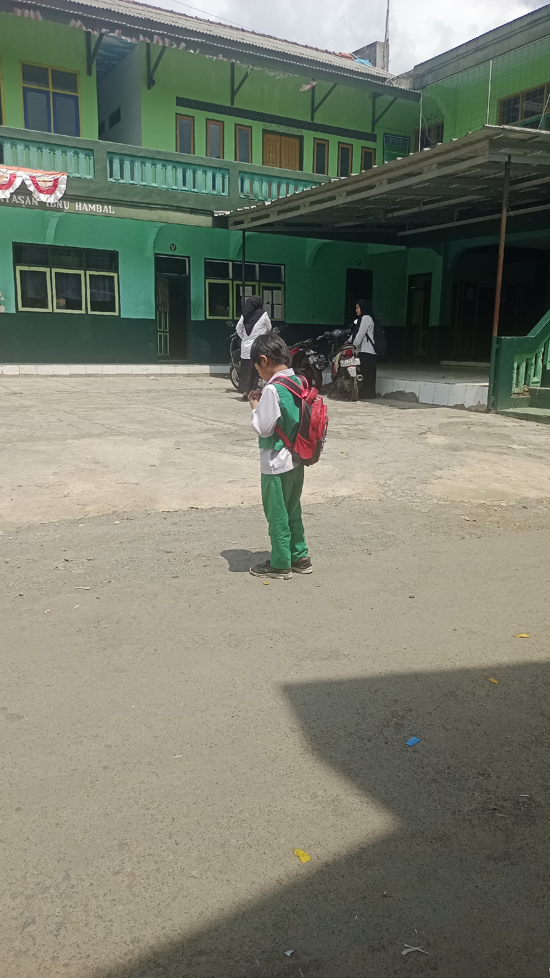 In Karyamandala, Indonesia, a young boy stands on a paved courtyard in front of a two-story green building with a covered walkway. The boy is wearing a white shirt, green pants, and a red backpack. He is looking down at his phone. Behind him, two women in white shirts and black hijabs are standing near motorcycles. The building has red and white banners hanging from its facade. The sky is cloudy.