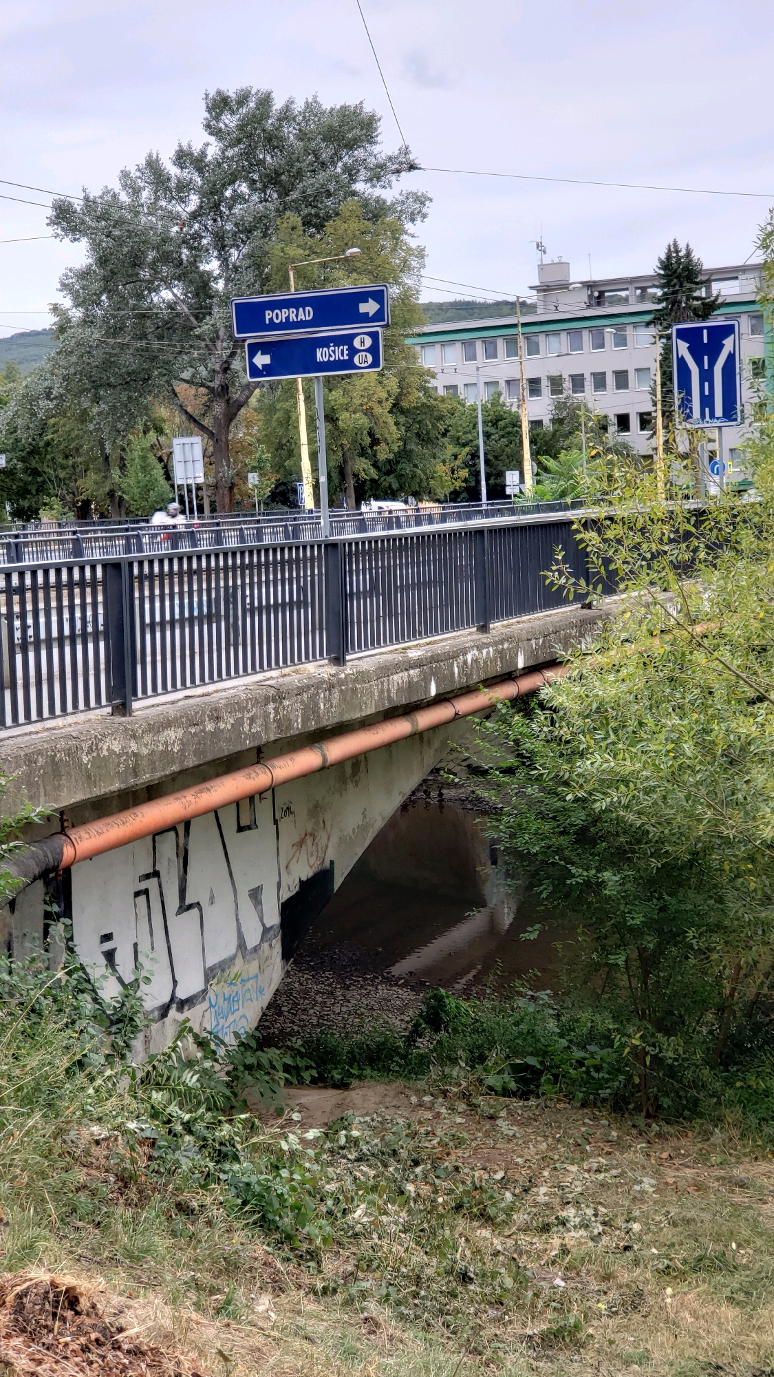 The image captures a view from under a bridge in the city of Prešov, Slovakia. The underside of the bridge is adorned with graffiti, with large white letters prominently displayed. An orange pipe runs along the exterior of the bridge's underside. Above the bridge, road signs indicate directions for "POPRAD" with an arrow pointing right, and "KOŠICE" with an arrow pointing left. A blue road sign with white markings showing an intersection and lane division is also visible to the right. The scene includes lush green vegetation and trees on either side, with a body of water flowing beneath the bridge. The sky is overcast. The bridge itself features a black metal railing.