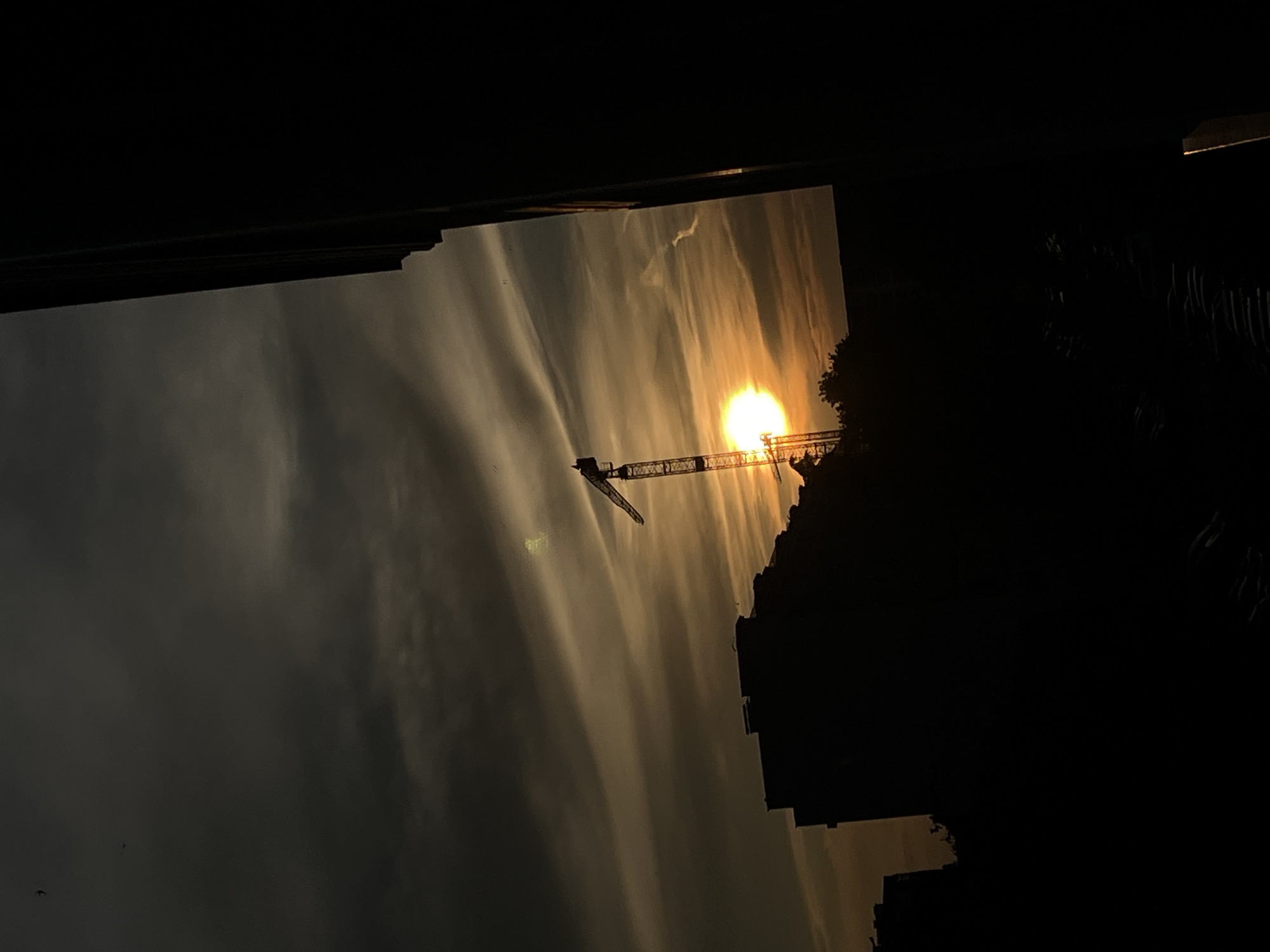 A silhouette of a construction crane and buildings is visible against a sunset sky. The sun, a bright orb of orange and yellow, is partially obscured by the crane and a building on the left. Wispy clouds streak across the sky, reflecting the warm hues of the sunset. The scene is dark, with the foreground and right side of the image dominated by deep shadows, suggesting a viewpoint from within a dark building or room looking out. The city of Burgas, Bulgaria, is indicated by the presence of a construction site near the coast, which often features cranes. The overall atmosphere is one of urban development against a dramatic natural backdrop.