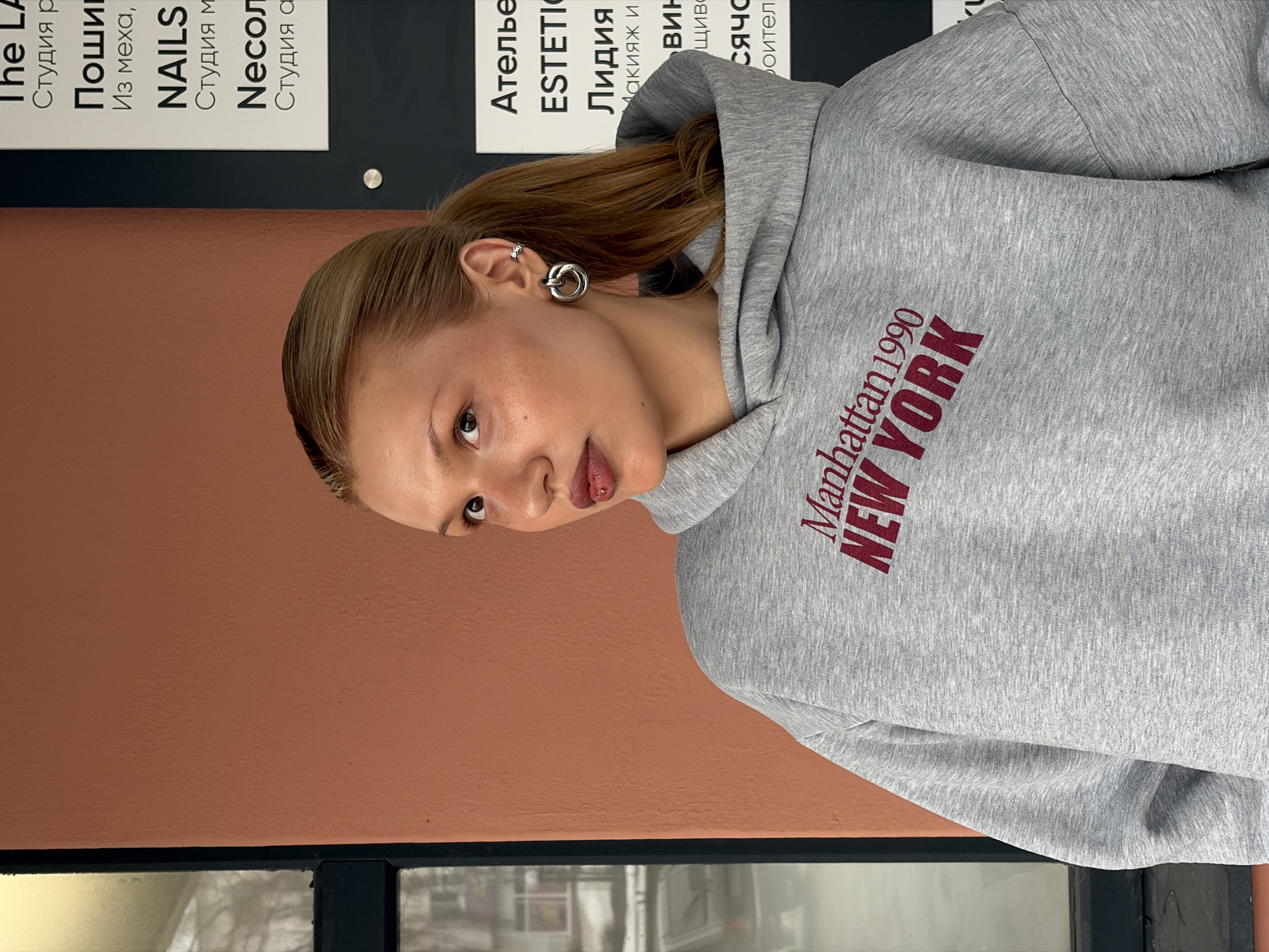 A fair-skinned young woman with light brown hair tied back is angled to the left, wearing a gray hoodie with "Manhattan 1990 NEW YORK" printed in maroon. She has multiple piercings in her left ear, including a double hoop earring. Her gaze is directed at the viewer.
She is situated next to a brown wall above a glass-paned window. Mounted on the wall is a sign with Russian text. Text visible on the sign includes the phrases "The LA," "NAILS," "ESTETIC," and "Лидия."
The location is identified in the city of Chaikovsky, Russia.