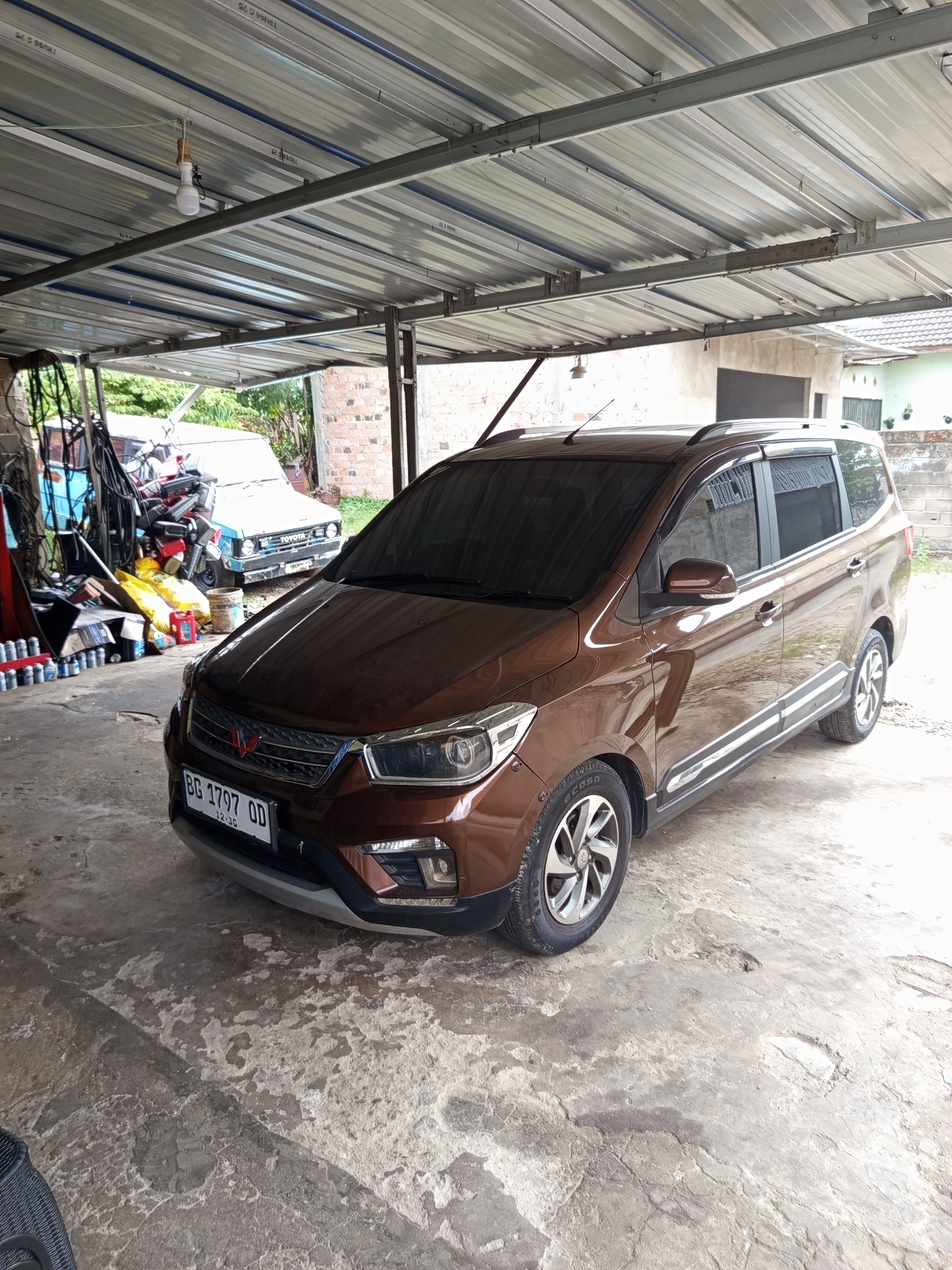The image depicts a brown Wuling Confero car parked under a metal canopy, likely a carport or garage, in Palembang, Indonesia. The car is positioned at an angle, with its front slightly towards the left of the frame. The license plate reads "BG 1797 OD". The ground is a rough, uneven concrete surface.
In the background, to the left, there is a cluttered area with various objects, including what appears to be a white Toyota car, motorcycles, tools, and containers. The area is partially open to an outdoor environment with greenery visible. To the right of the Wuling car, a light-colored building with a brick section is visible. The roof of the canopy is made of corrugated metal, supported by metal beams. A light bulb hangs from the ceiling. The overall impression is of a utilitarian space, possibly a residential or workshop area. There are no people visible in the image.