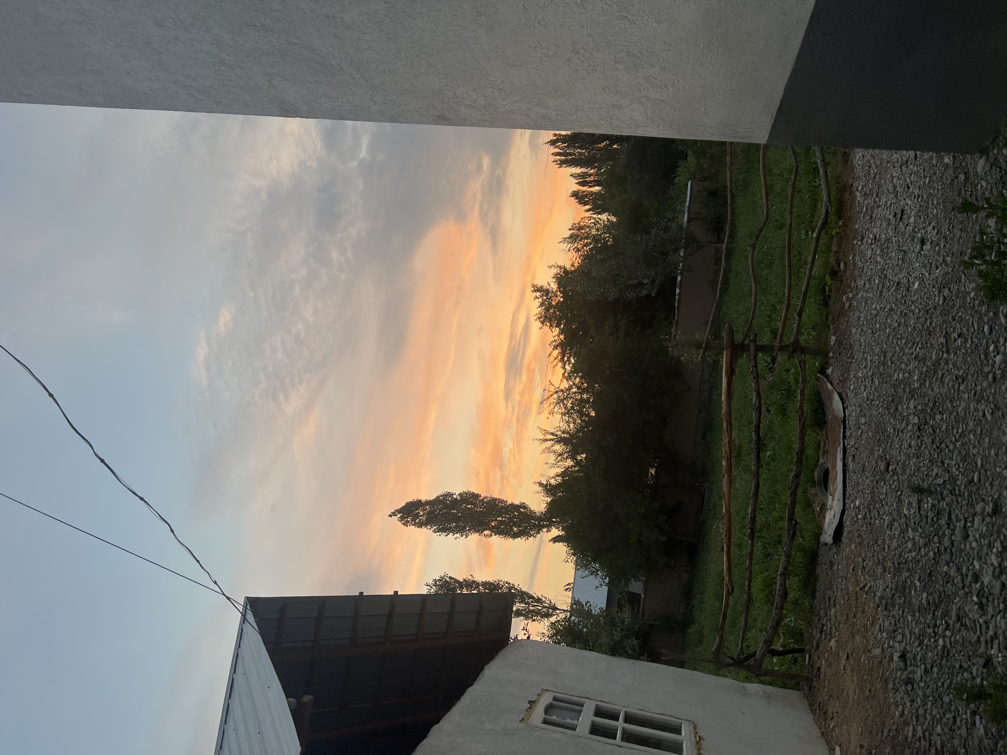 The image depicts a rural scene in Kara-Dıykan, Kyrgyzstan, during what appears to be sunset or early evening. The sky is filled with soft, warm colors, transitioning from pale grey to hues of peach and orange, suggesting the sun is low on the horizon. Wispy clouds streak across the sky.
In the foreground, the edge of a white building with a textured surface is visible at the top of the frame, and another building with a dark, corrugated metal roof is partially seen on the left. A rough, grey stone-covered ground extends to the right. A simple wooden fence, constructed from rough branches, borders a green grassy area. Several tall, slender trees are prominent in the middle ground, with dense foliage. In the distance, more trees form a dark line against the colorful sky. Power lines are also visible, strung across the sky. There is no discernible activity or human presence, giving the scene a quiet, still atmosphere. The overall mood is serene and peaceful, enhanced by the soft, natural light of dusk.
