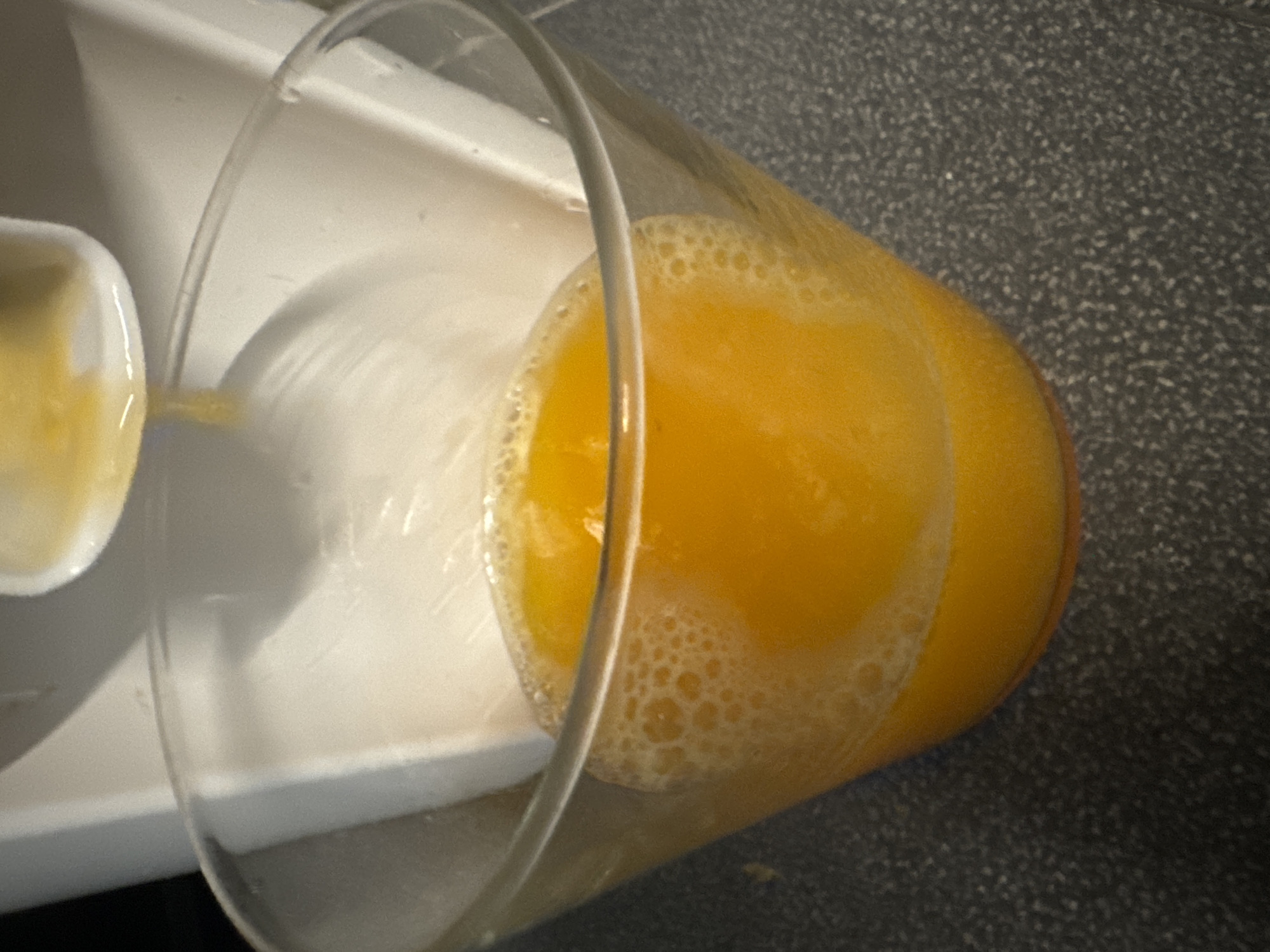This image shows a close-up view of a clear glass filled with freshly squeezed orange juice. The juice is a vibrant orange color and has a frothy top layer with small bubbles. The glass is placed on a dark, speckled countertop. In the background, a white appliance, likely a juicer, is partially visible, with more orange juice dripping from its spout into the glass. The lighting is somewhat dim, highlighting the texture and color of the juice. The scene does not provide any information or context that would allow for the determination of its location in the city of Murcia, Spain.