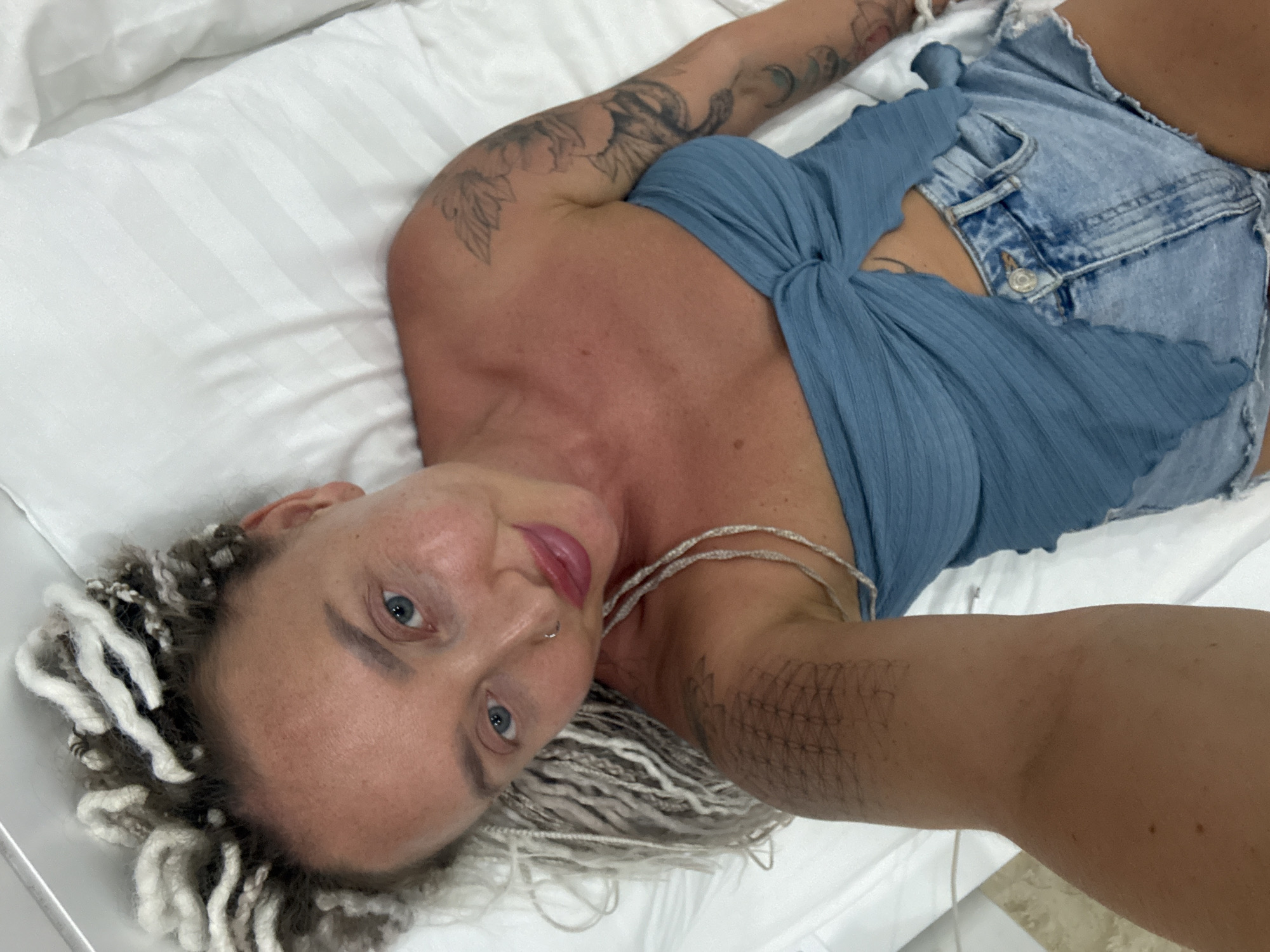 A woman is lying on a white striped surface, possibly a bed or couch. She has blonde hair with streaks of grey, styled into braids. She is wearing a blue, ribbed crop top and denim shorts. Her left arm is visible, with tattoos on the shoulder and forearm. She is looking towards the camera with blue eyes and has a nose ring and a necklace made of silver beads. Her lips are painted pink. The lighting suggests it is daytime, and the indoor setting implies a private space. There is no visible text in the image.