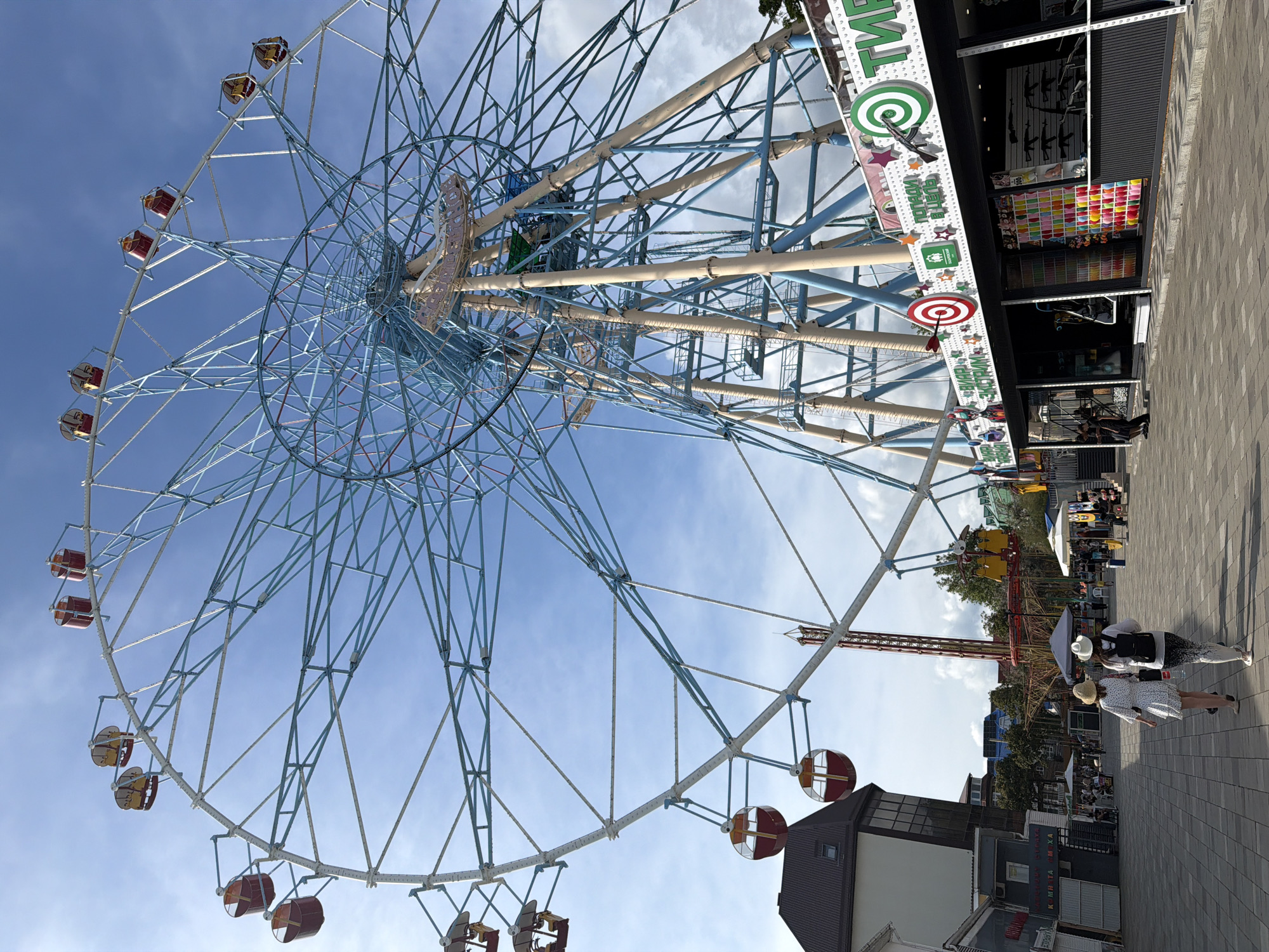 The image depicts an amusement park setting, likely in Sochi, Russia, with a prominent Ferris wheel as the central element. The Ferris wheel is large and blue, with red and yellow gondolas. In the foreground, two women are walking away from the camera on a paved walkway. To the right of the Ferris wheel, there is a shooting gallery attraction with targets and prizes displayed. Buildings typical of a resort town, including a dark-roofed building with windows and several smaller structures, are visible to the left. The sky is partly cloudy and blue. The atmosphere suggests a sunny day in an entertainment district.