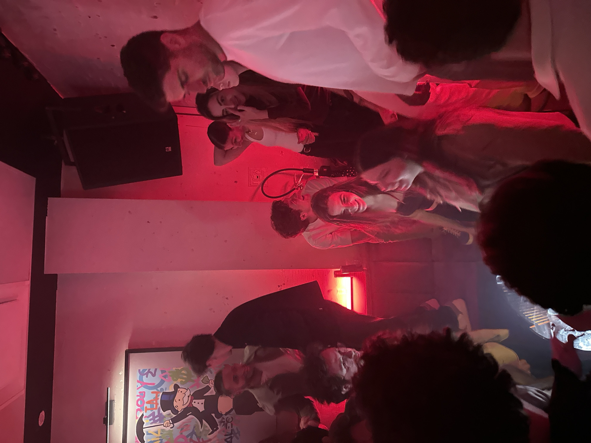 A dimly lit room, likely a bar or club in Mexico City, Mexico, features a gathering of people. The lighting is predominantly red, creating a vibrant atmosphere. There is a speaker positioned on the left. A poster with a cartoon is affixed to the lower left wall. A man wears a white shirt; other attendees are dressed in darker colors. People are standing or sitting in groups. Interactions are unclear due to the perspective and lighting. The environment appears crowded and social.