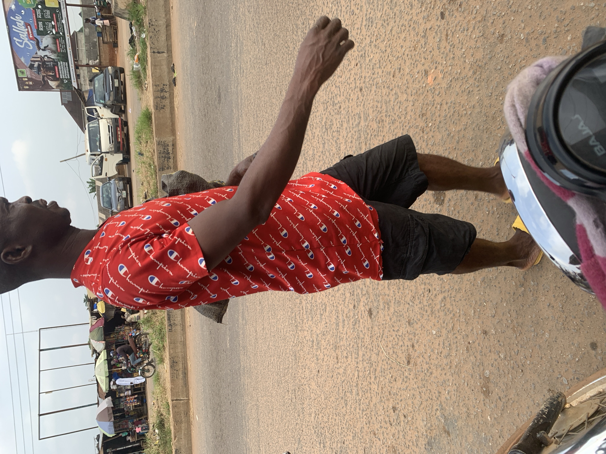 A full shot shows a dark-skinned man walking across a dusty, paved road in the city of Shagamu, Nigeria. He wears a red Champion t-shirt and black shorts, with yellow sandals. He carries a dark bag over his left shoulder. The background features a row of shops with colorful awnings, several vehicles including vans and cars, and a large billboard advertising land plots. The man is walking away from the camera, his right arm extended. The scene is an outdoor market or roadside area.