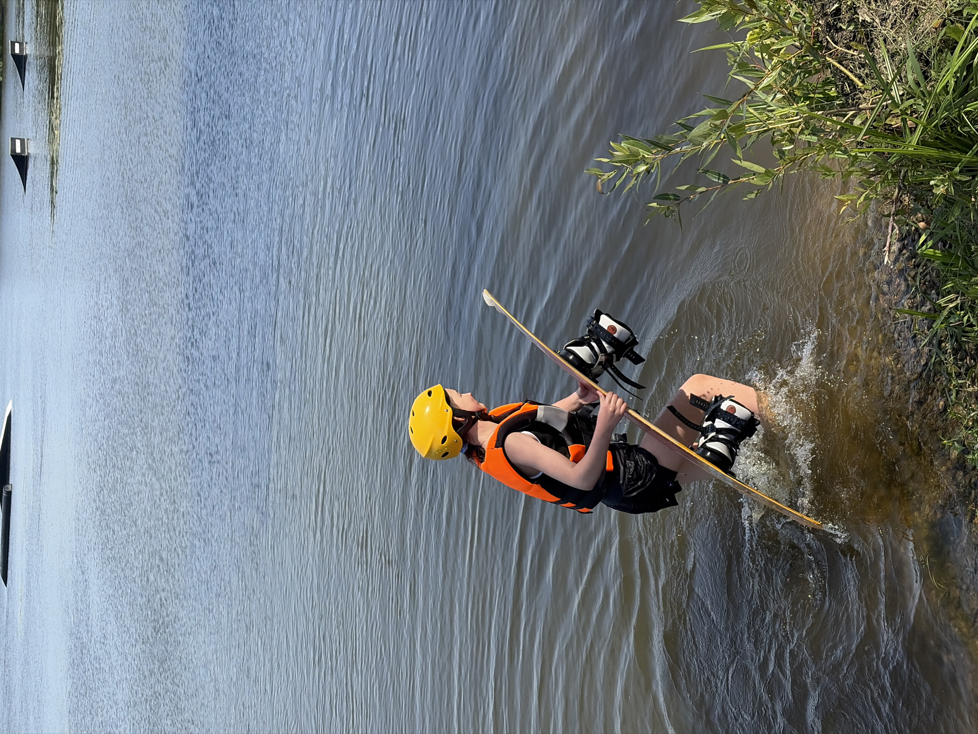 A person wearing a yellow helmet and an orange life vest is standing in shallow water, holding a wakeboard. The wakeboard is positioned vertically in the water, with bindings visible. The person is wearing black shorts. The background shows a large body of water with several ramps or obstacles in the distance. The foreground features the edge of a grassy bank with some vegetation. The image appears to be taken on a sunny day. The location is the city of Astrashitskagaradokski rural council, Belarus.